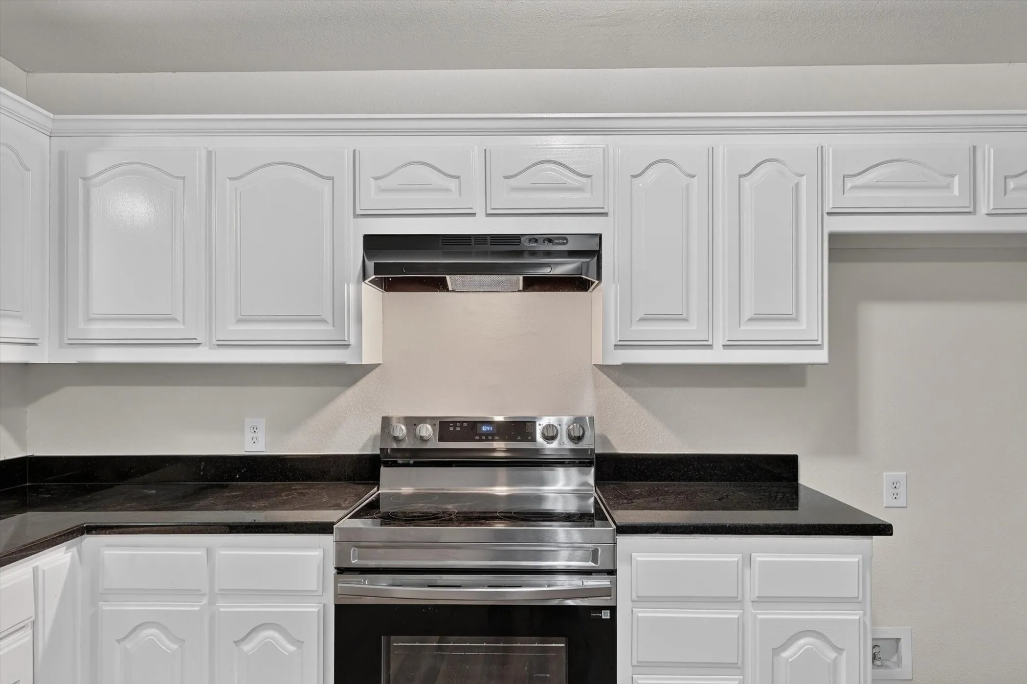 Kitchen featuring white cabinetry with arch panel doors and crown molding, dark stone countertops, and a stainless steel electric range with an overhead vent