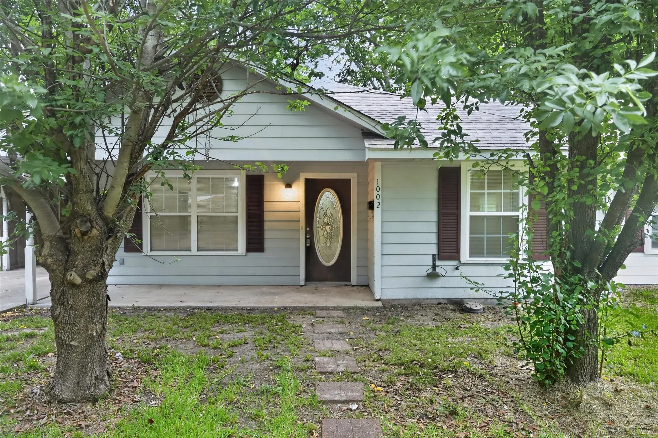 Light blue siding exterior featuring a dark wood entry door with an oval glass insert, two front windows with dark red shutters, and a covered concrete porch