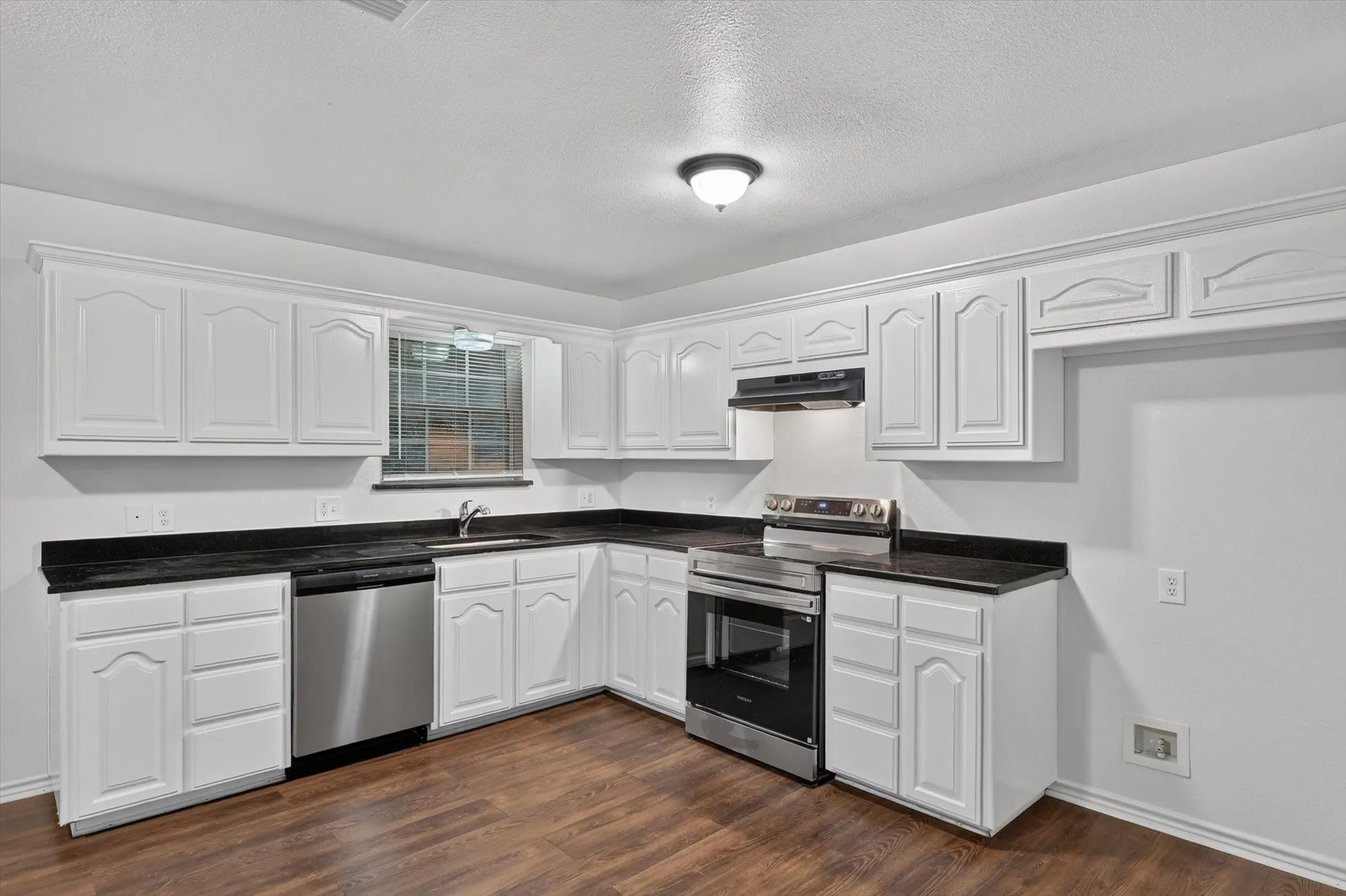 Kitchen featuring white cabinetry, dark countertops, a stainless steel dishwasher, a stainless steel range with overhead ventilation, and wood-finish flooring