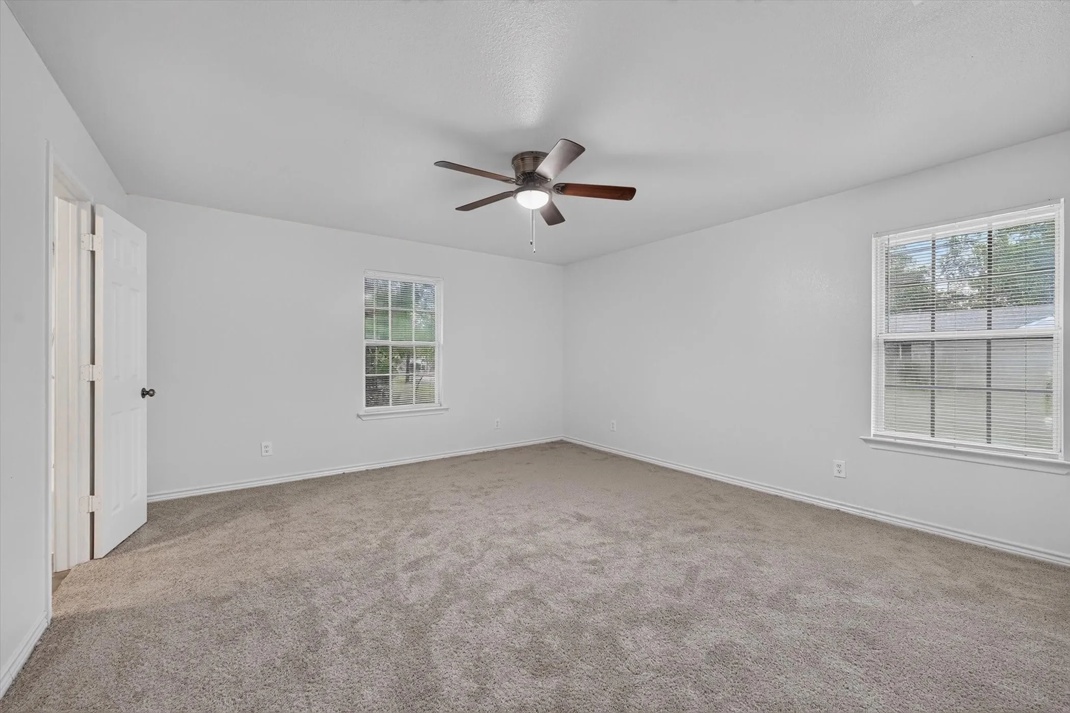 Spacious room featuring neutral carpeting, two windows with blinds, a ceiling fan with integrated lighting, and a white paneled door