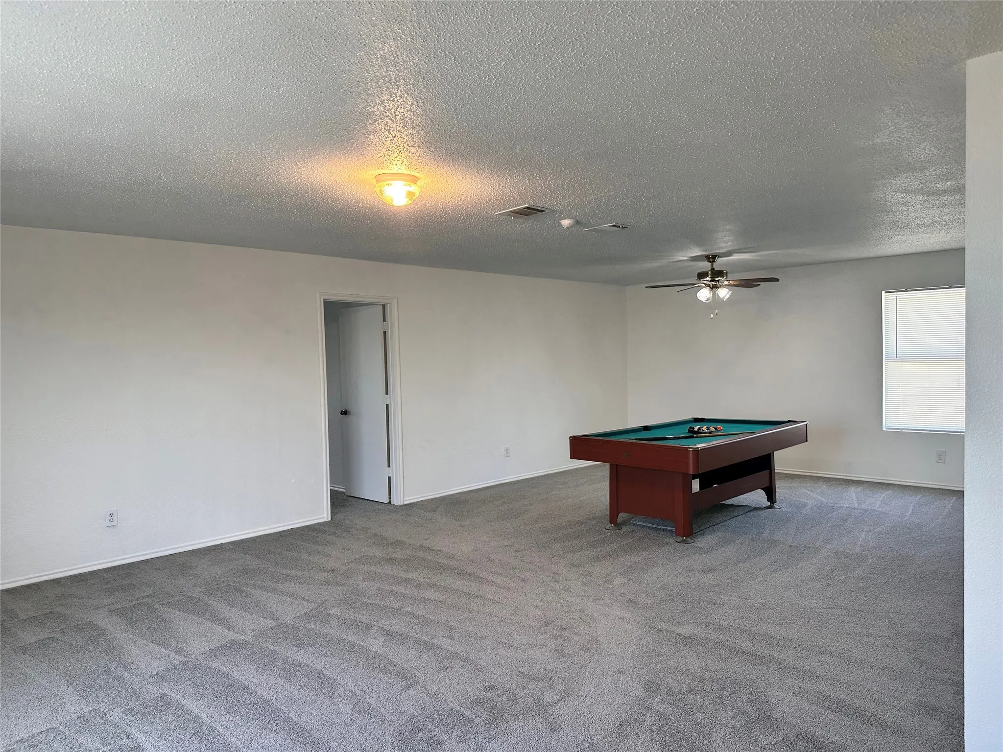 Spacious Game room Area with gray patterned carpeting, white walls, ceiling fan, and a window with blinds