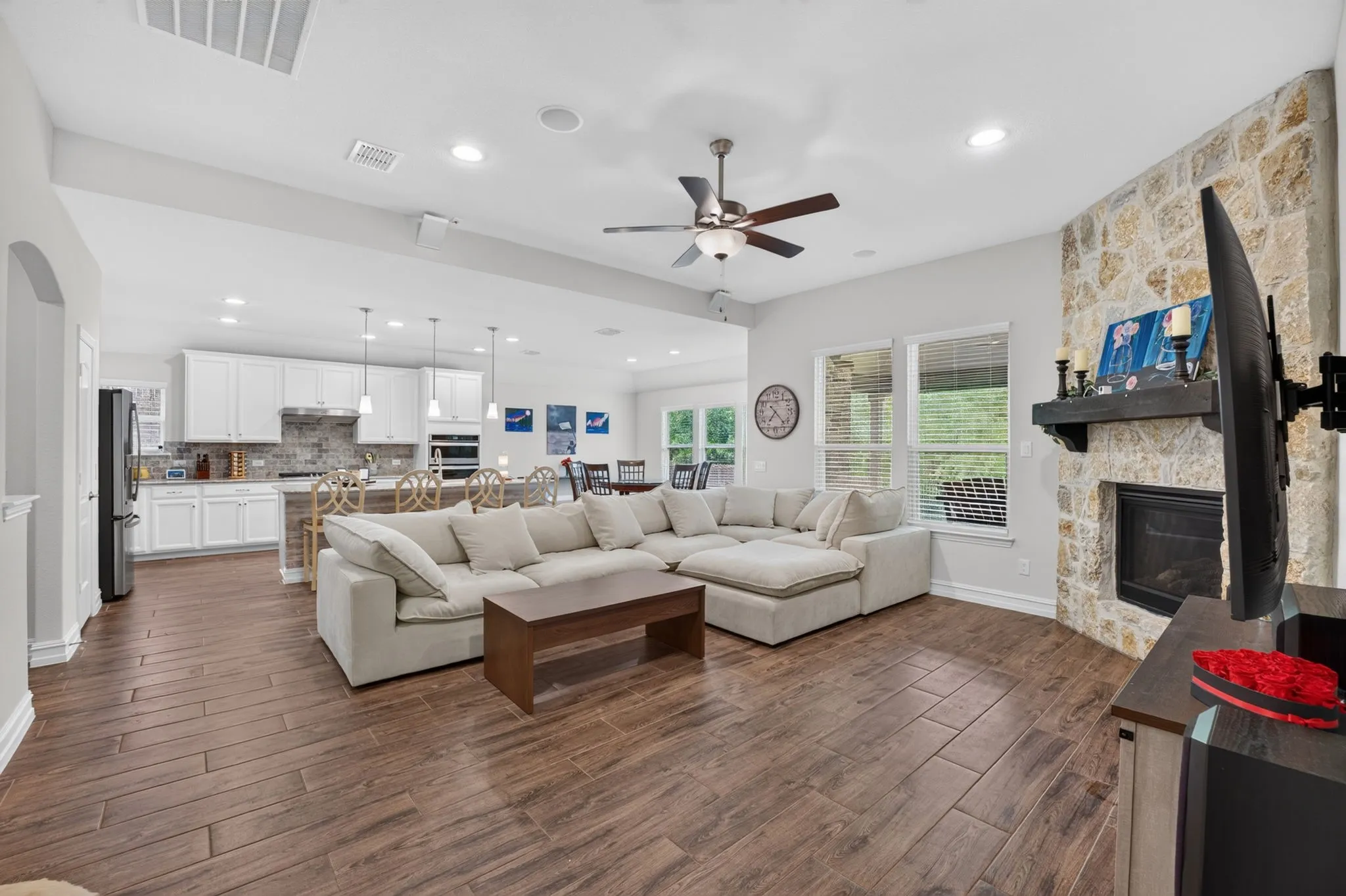 Open-concept living area featuring wood-finish flooring, a stone-faced fireplace with a dark wood mantel, and a ceiling fan