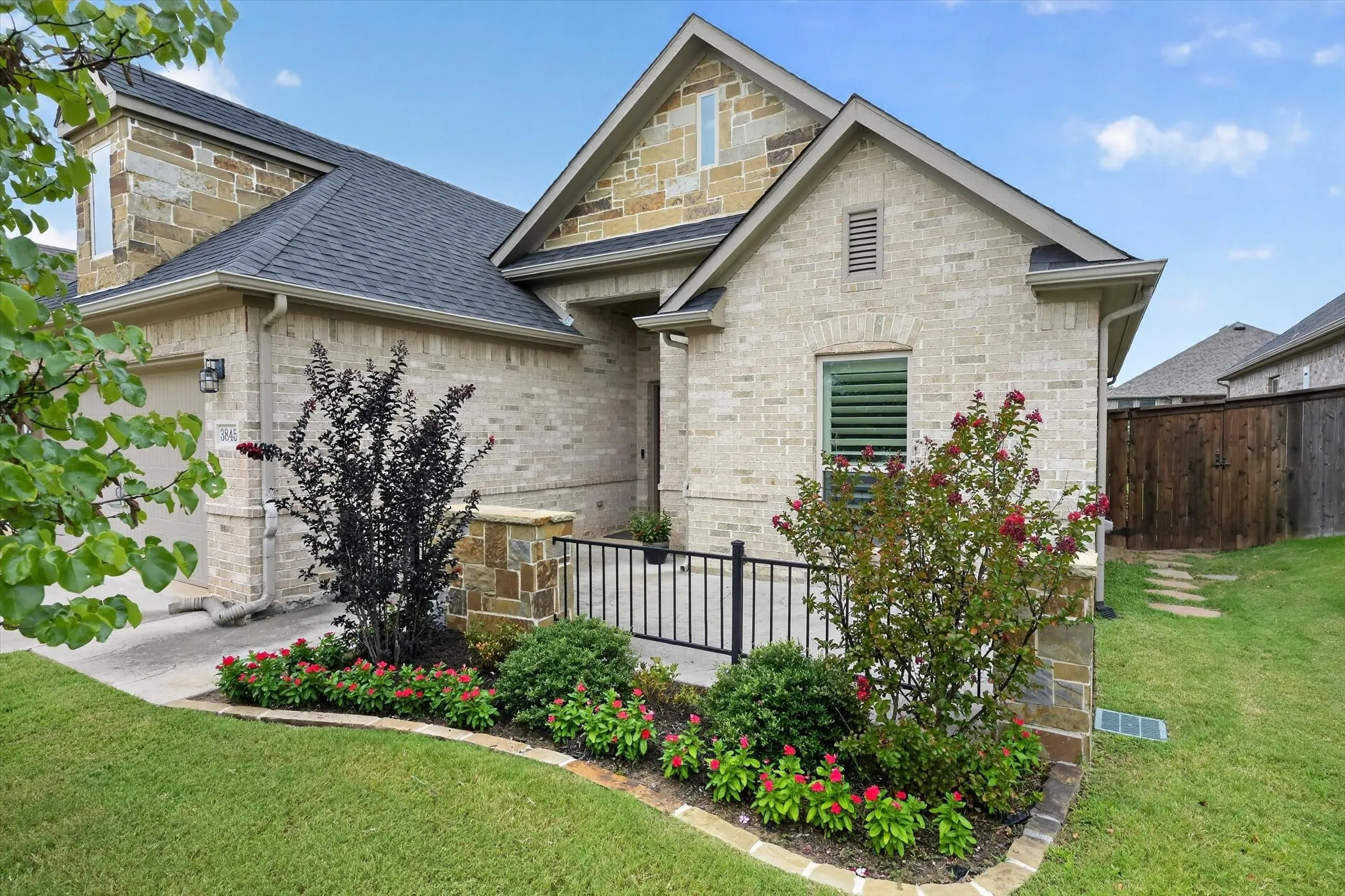Exterior featuring a blend of light-toned brick and natural stone accents, a dark shingle roof, a paved driveway, and meticulously landscaped flower beds