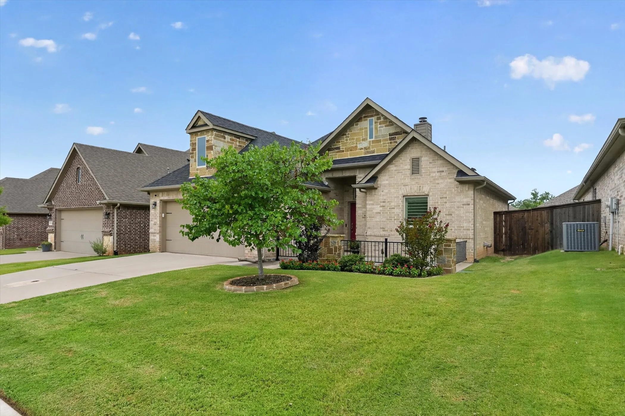 Brick and stone exterior featuring a covered front porch with iron railing, two-car garage, and landscaped front yard with a mature tree