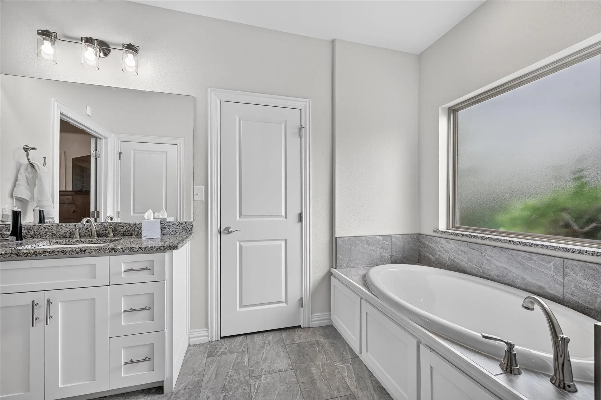 Bathroom featuring a built-in soaking tub, a large window with privacy glass, a vanity with a granite-style countertop, a chrome faucet, and gray tile flooring