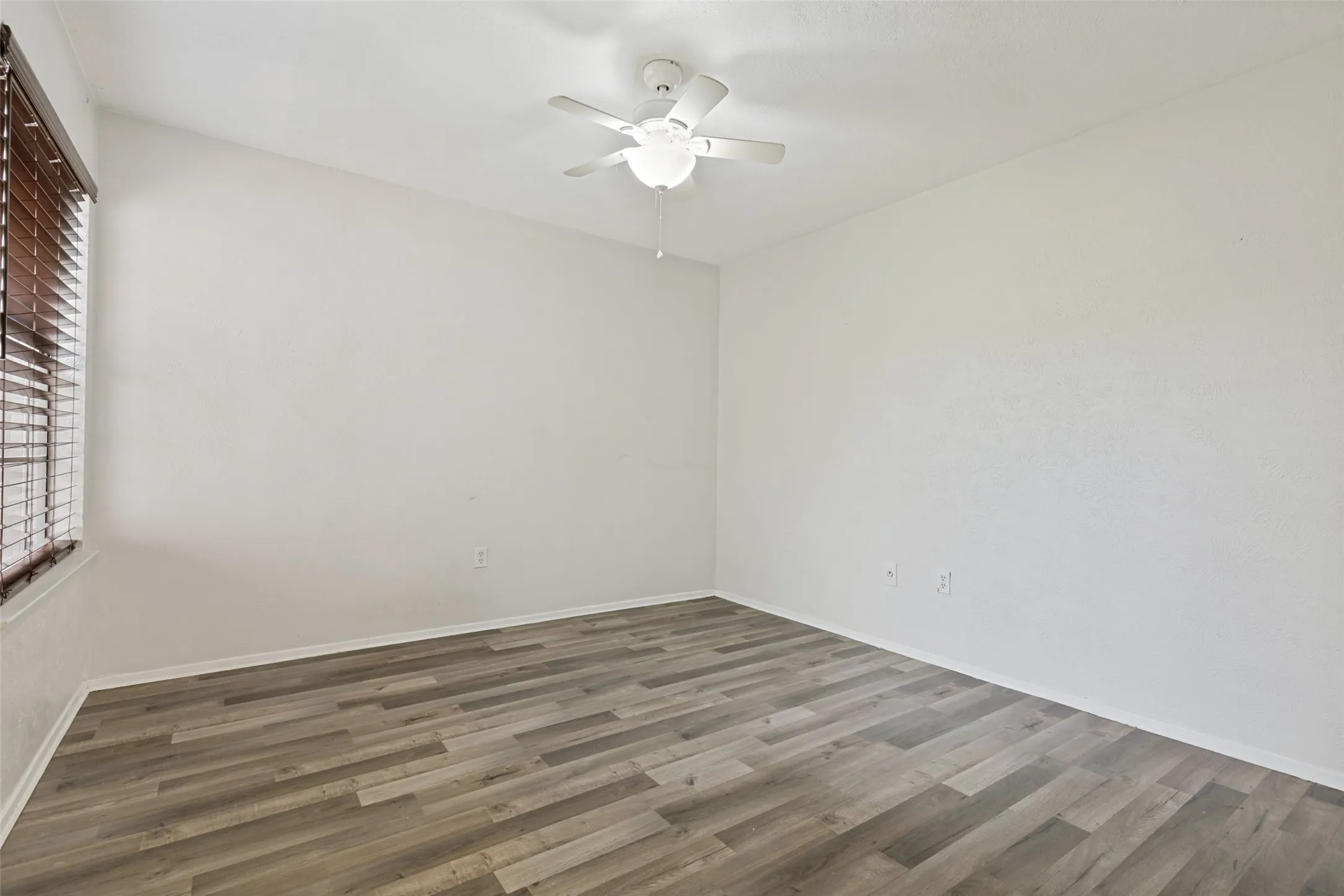 Spacious room featuring wood-finish flooring, white baseboards, and a ceiling fan with integrated lighting