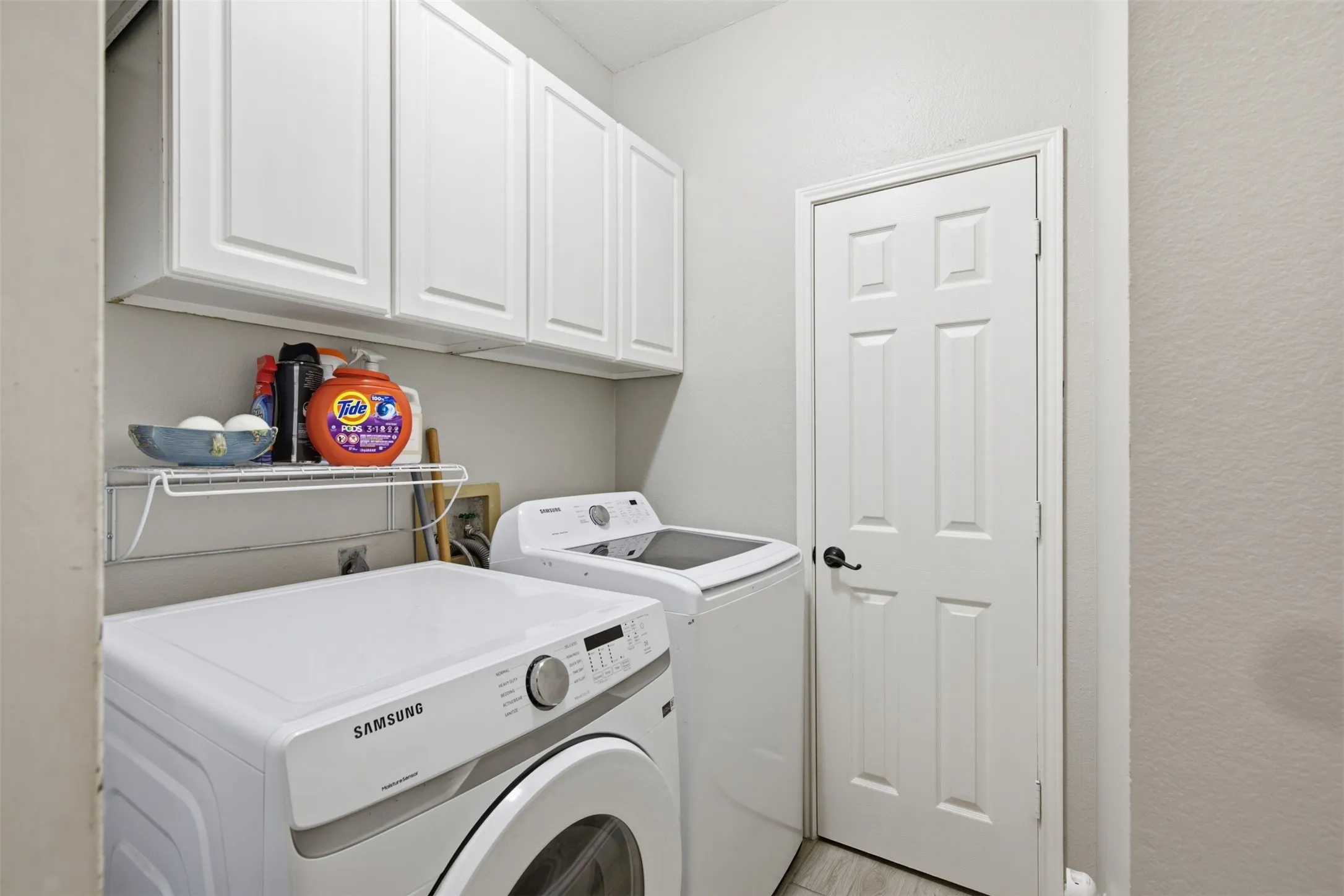 Dedicated utility space featuring white cabinetry, a white wire shelf, a panel door with black hardware, and light-toned flooring