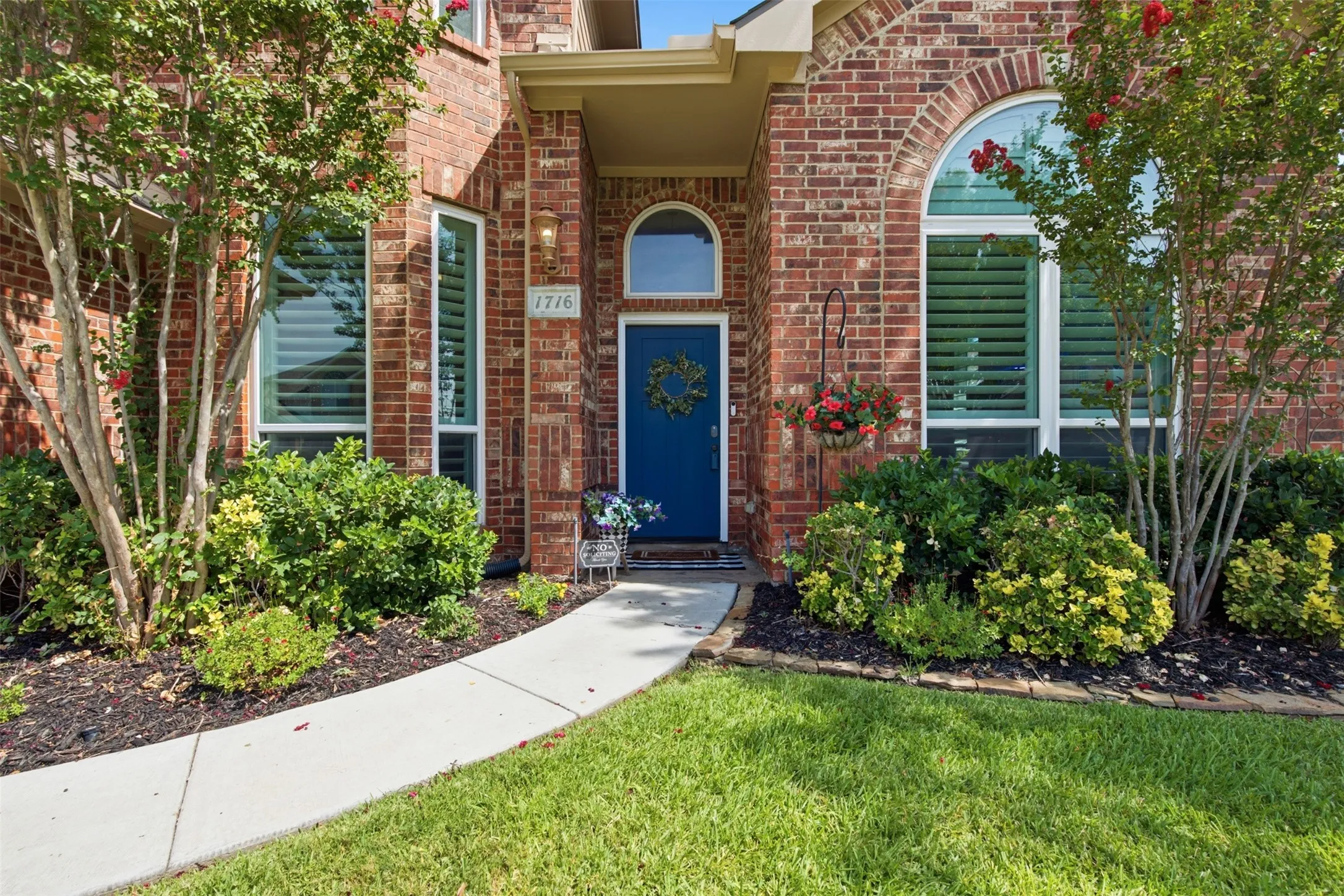 Brick exterior featuring a blue entry door with transom window, arched window with plantation shutters, front entry light fixture, and manicured landscaping with a concrete pathway