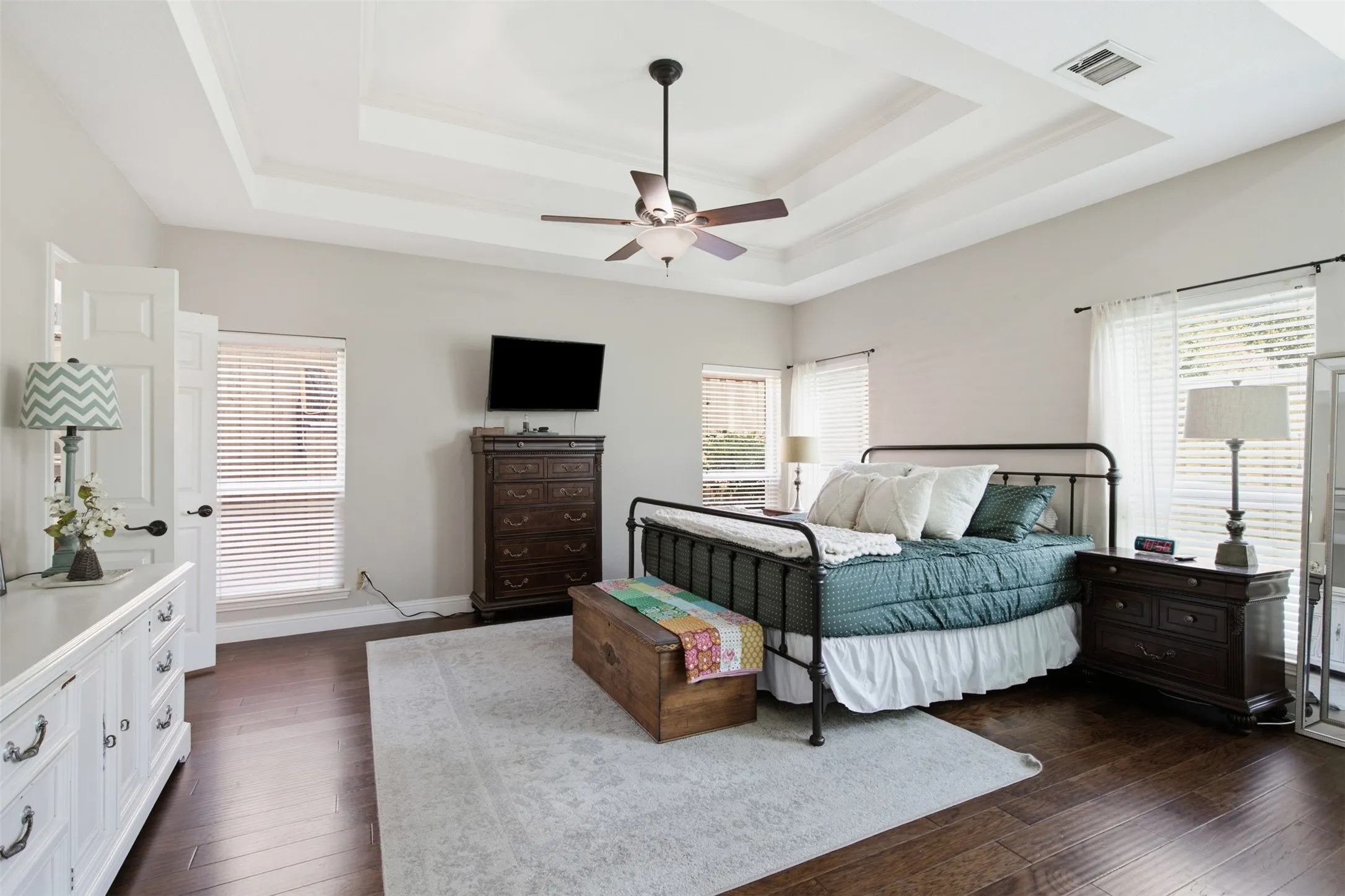 Spacious room featuring rich wood-finish flooring, a tiered tray ceiling with a ceiling fan, and multiple windows with horizontal blinds