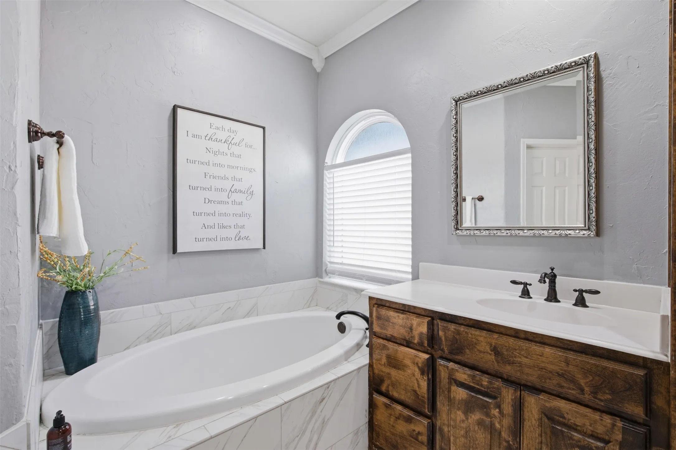 Soaking tub with surrounding marble-finish tile, arched window with privacy blinds, and crown molding