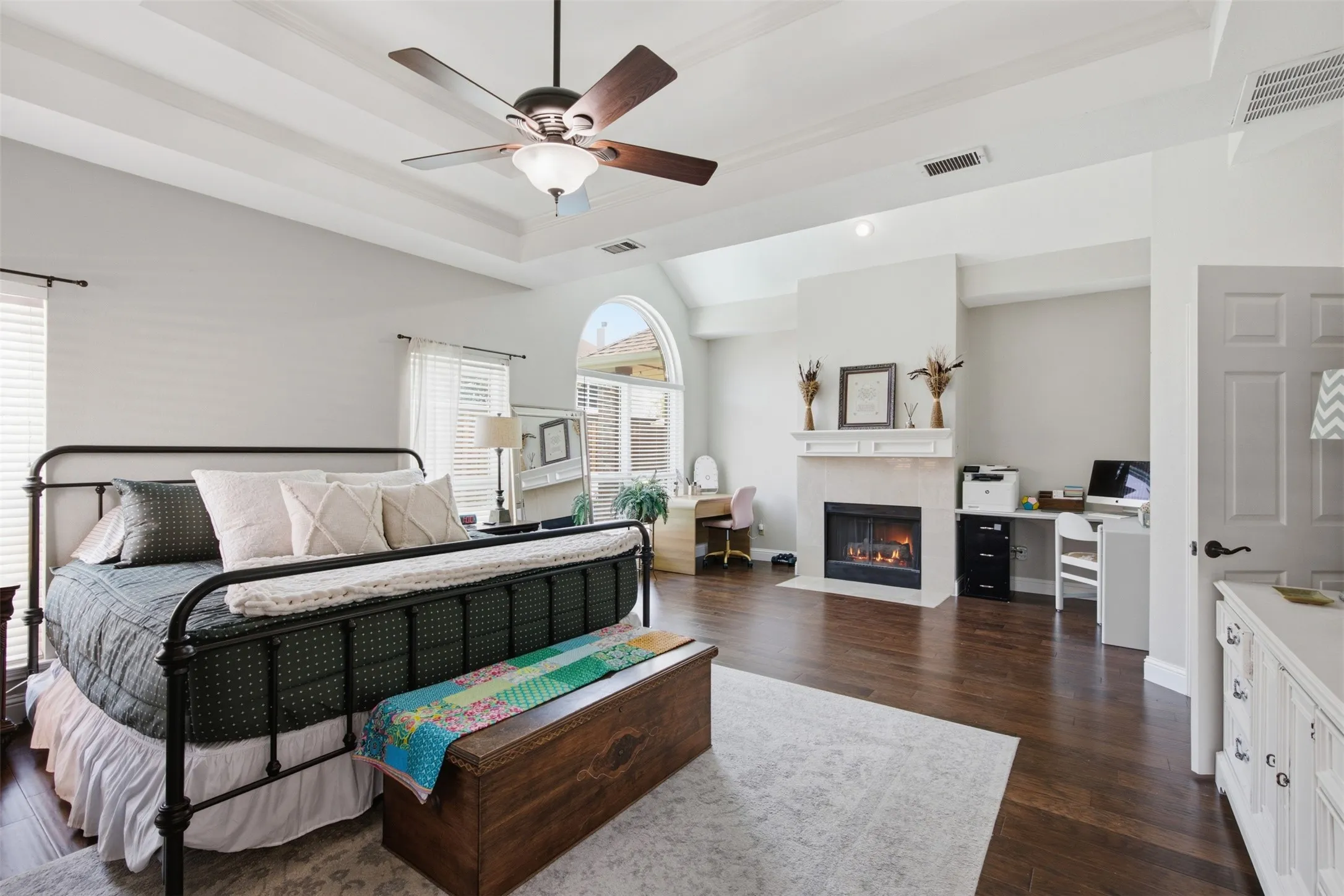 Spacious room featuring rich wood-finish flooring, a tray ceiling with a ceiling fan, and a fireplace with a light-colored surround