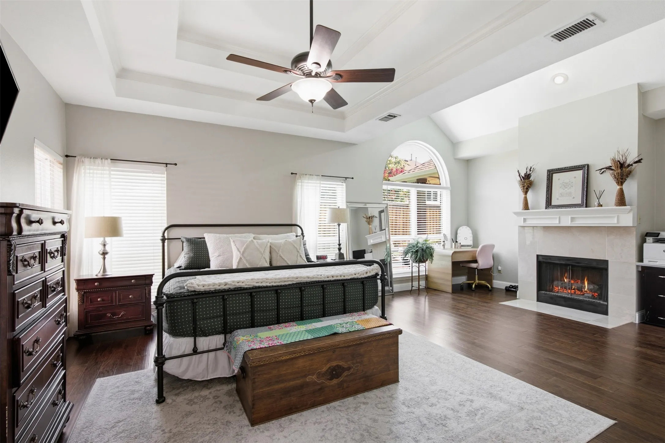 Tray ceiling with ceiling fan, wood-finish flooring, arched window, fireplace with white mantel, and vaulted ceiling