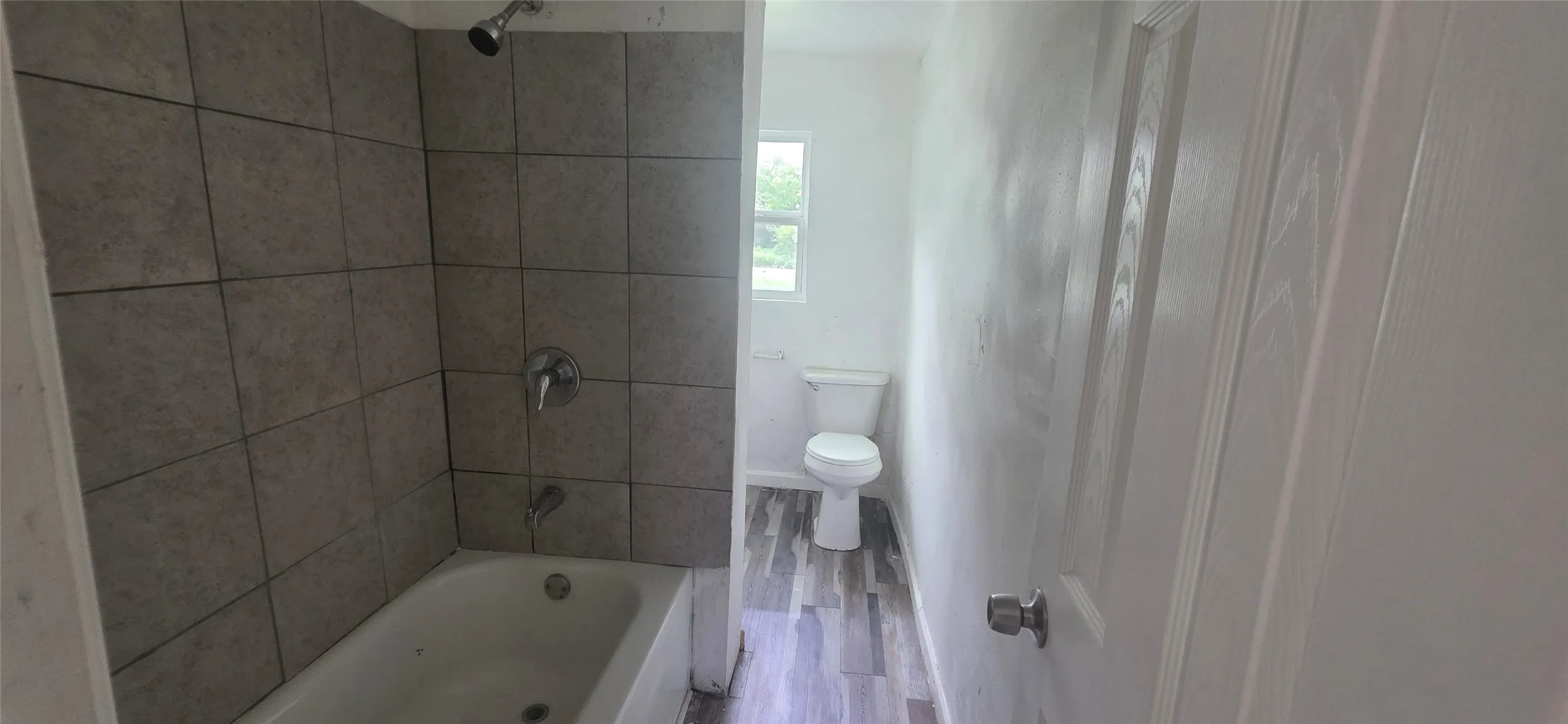 Bathroom featuring a white bathtub with a tiled shower surround, chrome fixtures, a window, a white toilet, and wood-finish flooring