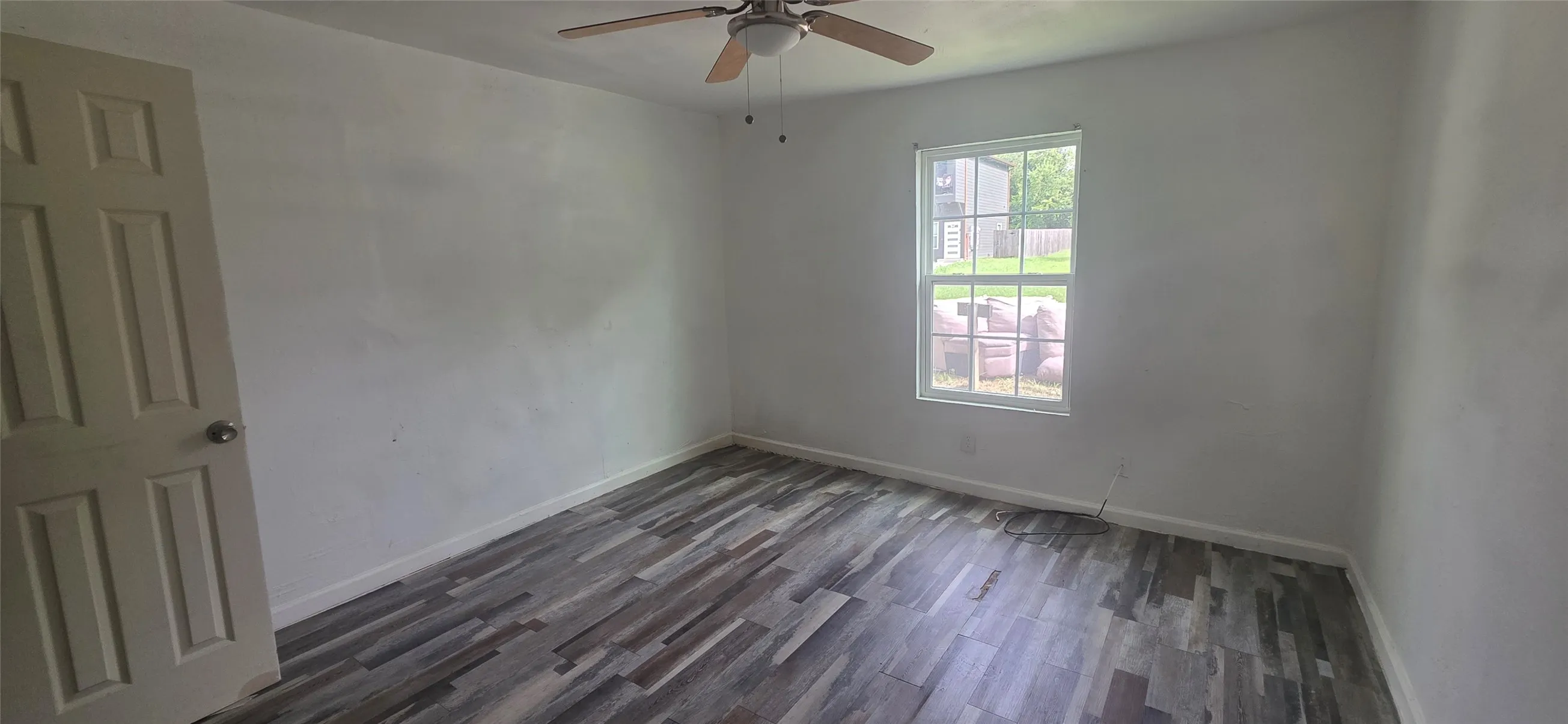 Room featuring multi-tone wood-finish flooring, a ceiling fan with light fixture, and a large window providing ample natural light