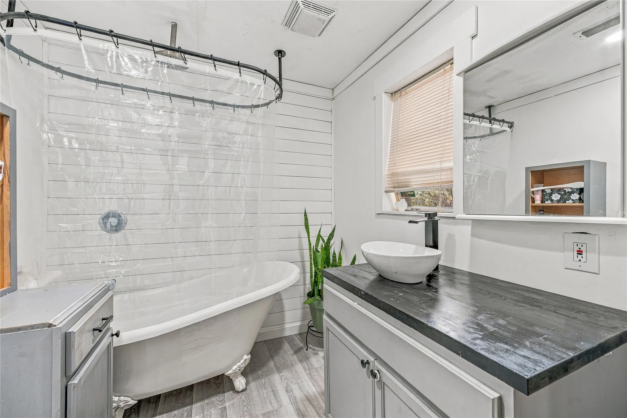 Freestanding clawfoot tub with an overhead shower, shiplap accent wall, wood-finish flooring, and a modern vanity with a vessel sink