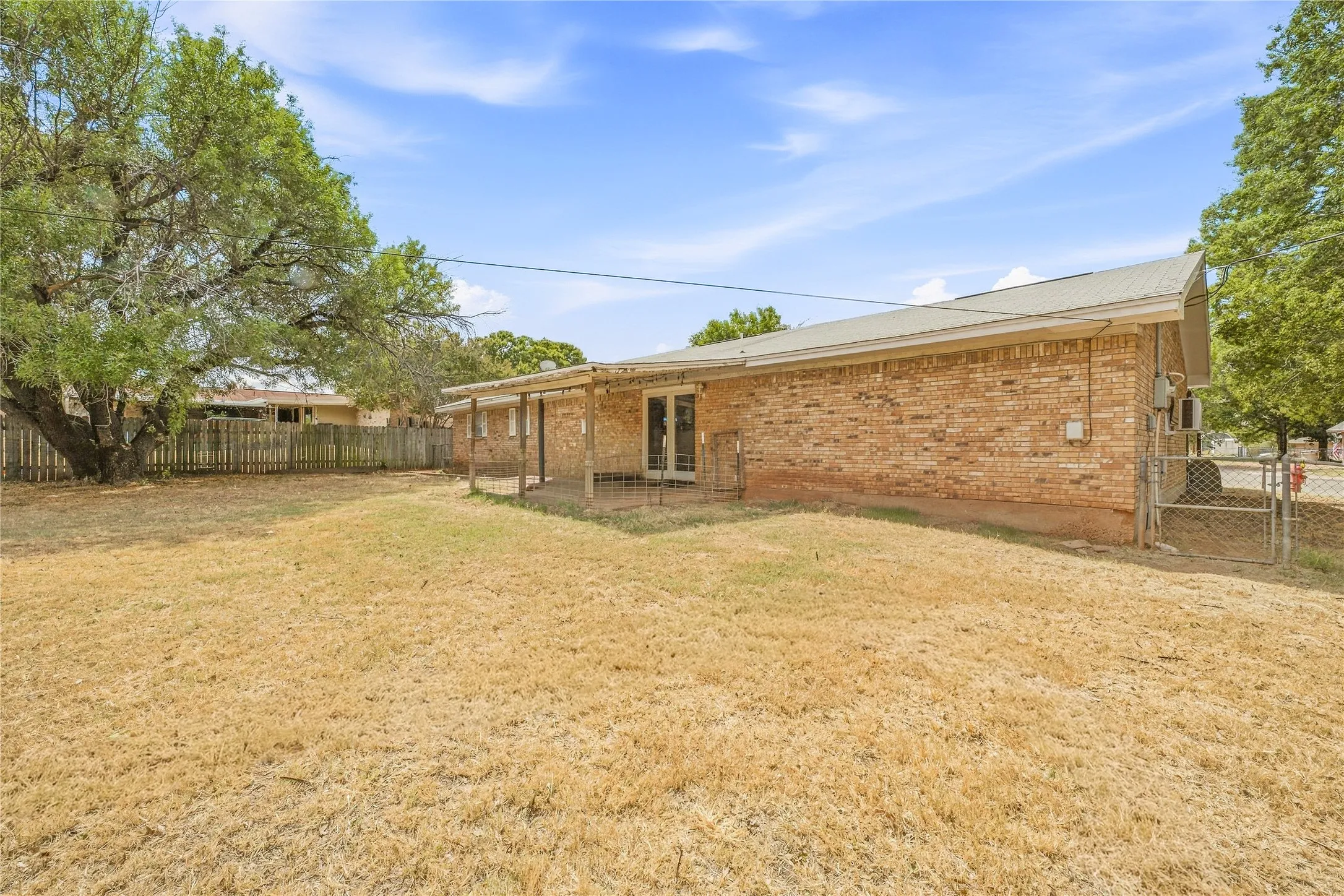 Brick exterior with a covered patio, a fenced yard, mature trees, and a chain-link gate