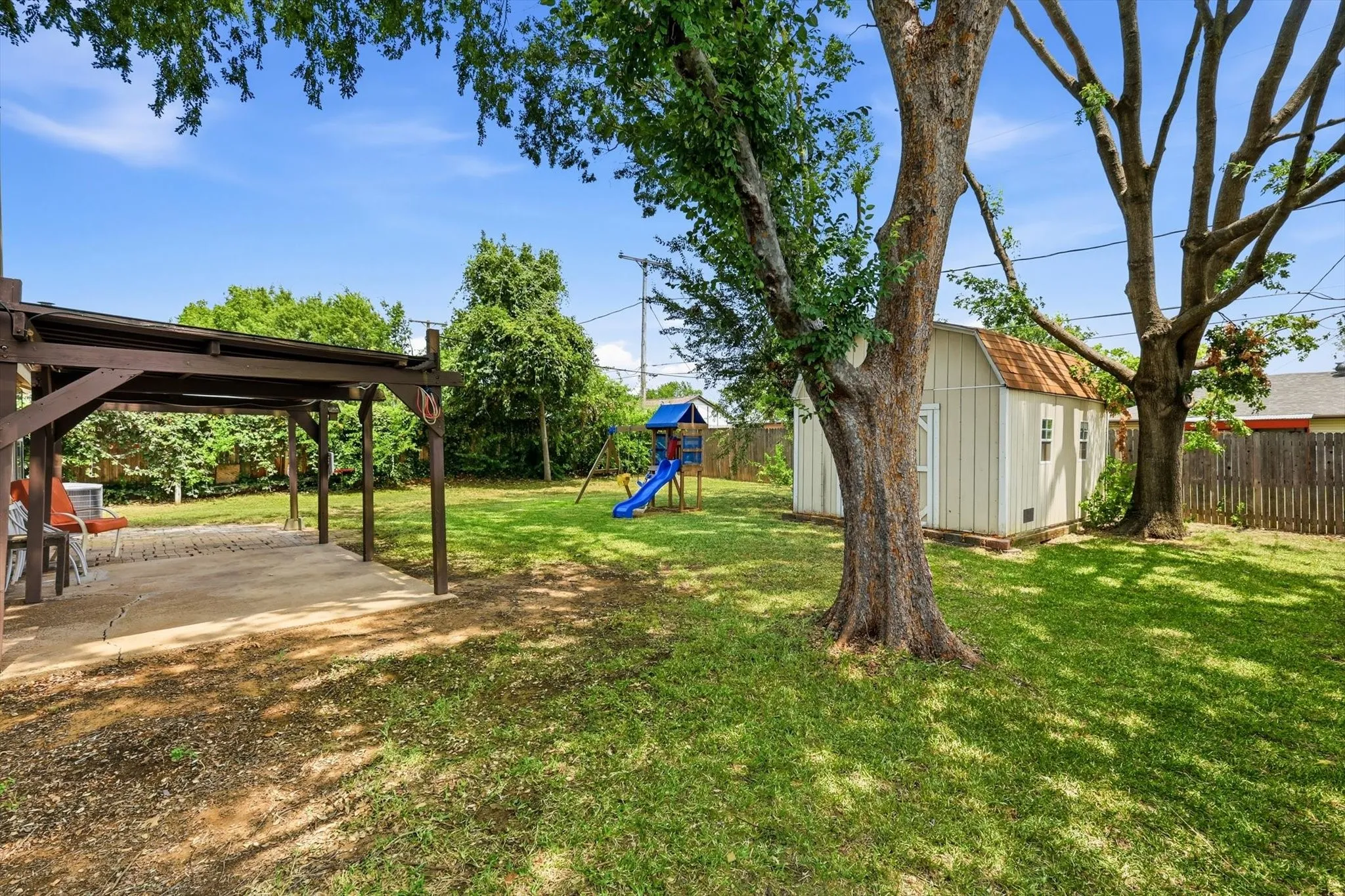 Expansive backyard featuring a large tree, a covered patio area with a concrete slab, a separate storage shed, and a privacy fence