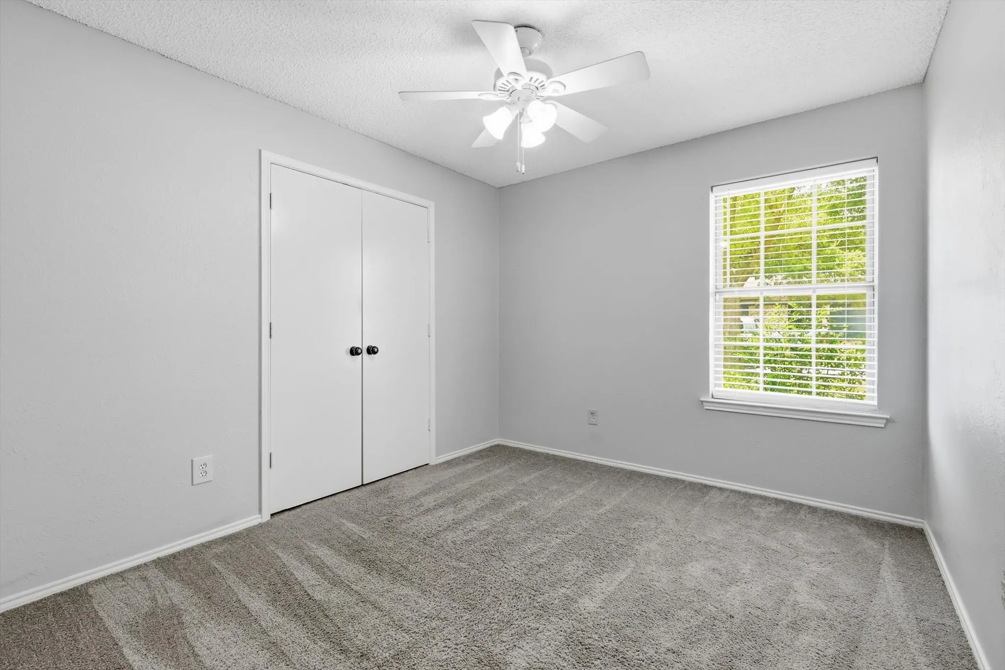 Neutral gray walls complemented by gray patterned carpeting, a ceiling fan with light fixture, white double-door closet with black hardware, and a window with horizontal blinds