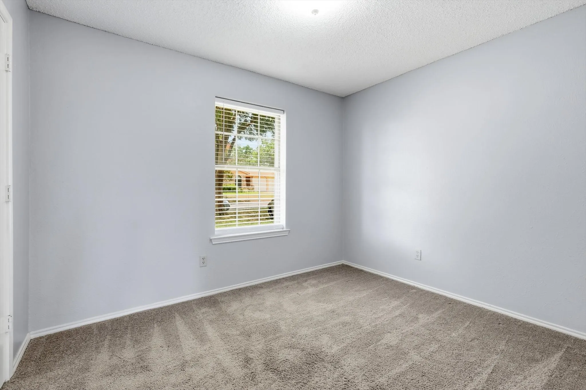 Bright room featuring a single pane window with horizontal blinds, light gray wall paint, white trim, and textured gray carpeting