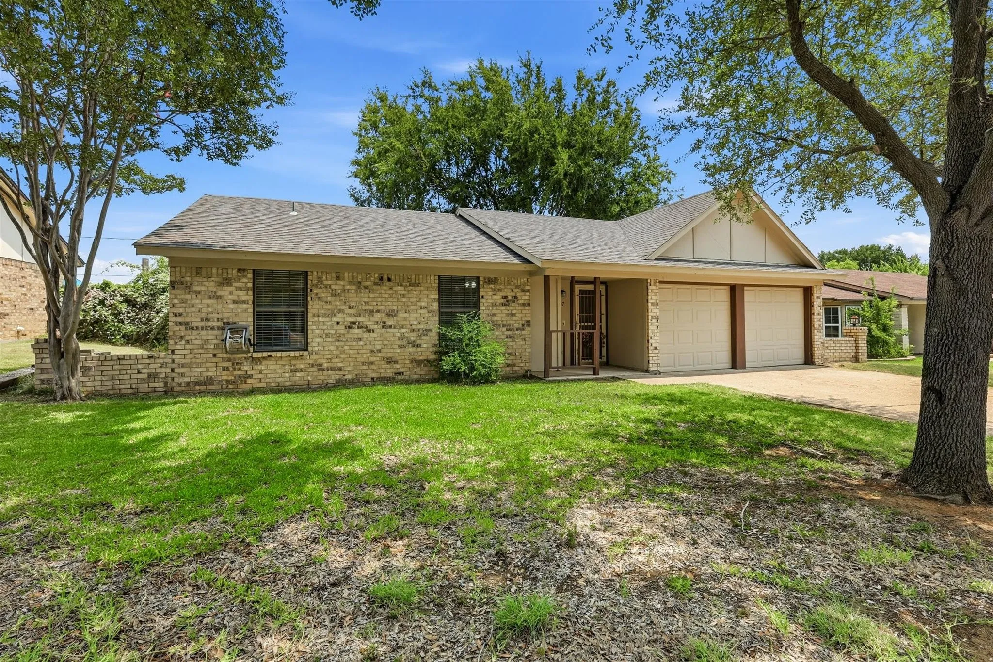 Brick exterior home featuring a two-car garage, mature front yard trees, and a covered entry