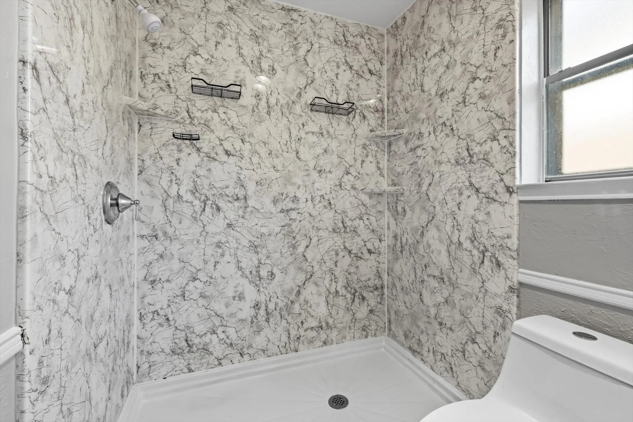 Shower enclosure featuring a white and gray marbled pattern, chrome fixtures, and a corner shelf