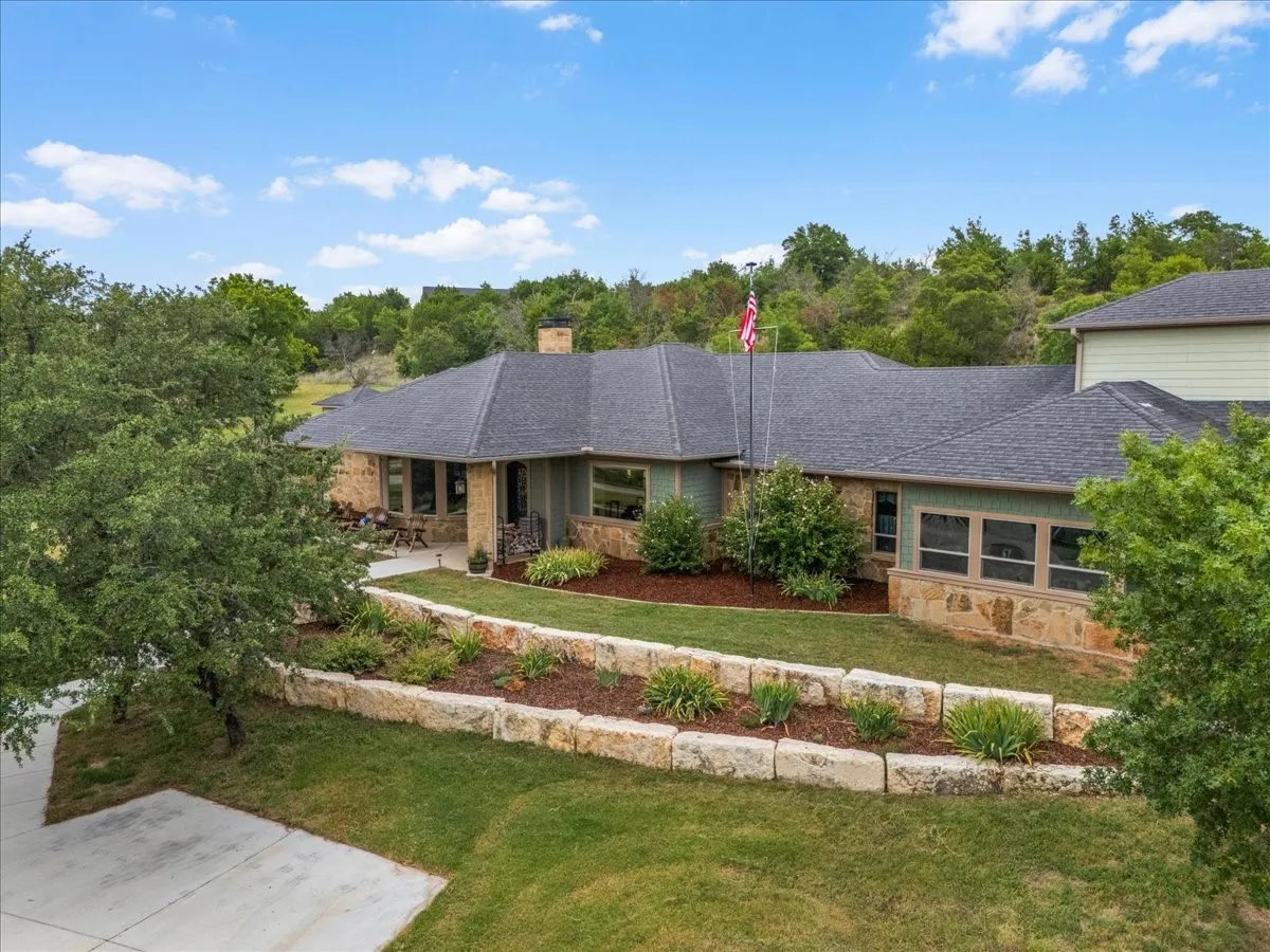 Expansive front exterior showcasing a stone and siding facade, a dark shingle roof, a concrete driveway, and tiered landscaping with natural stone retaining walls