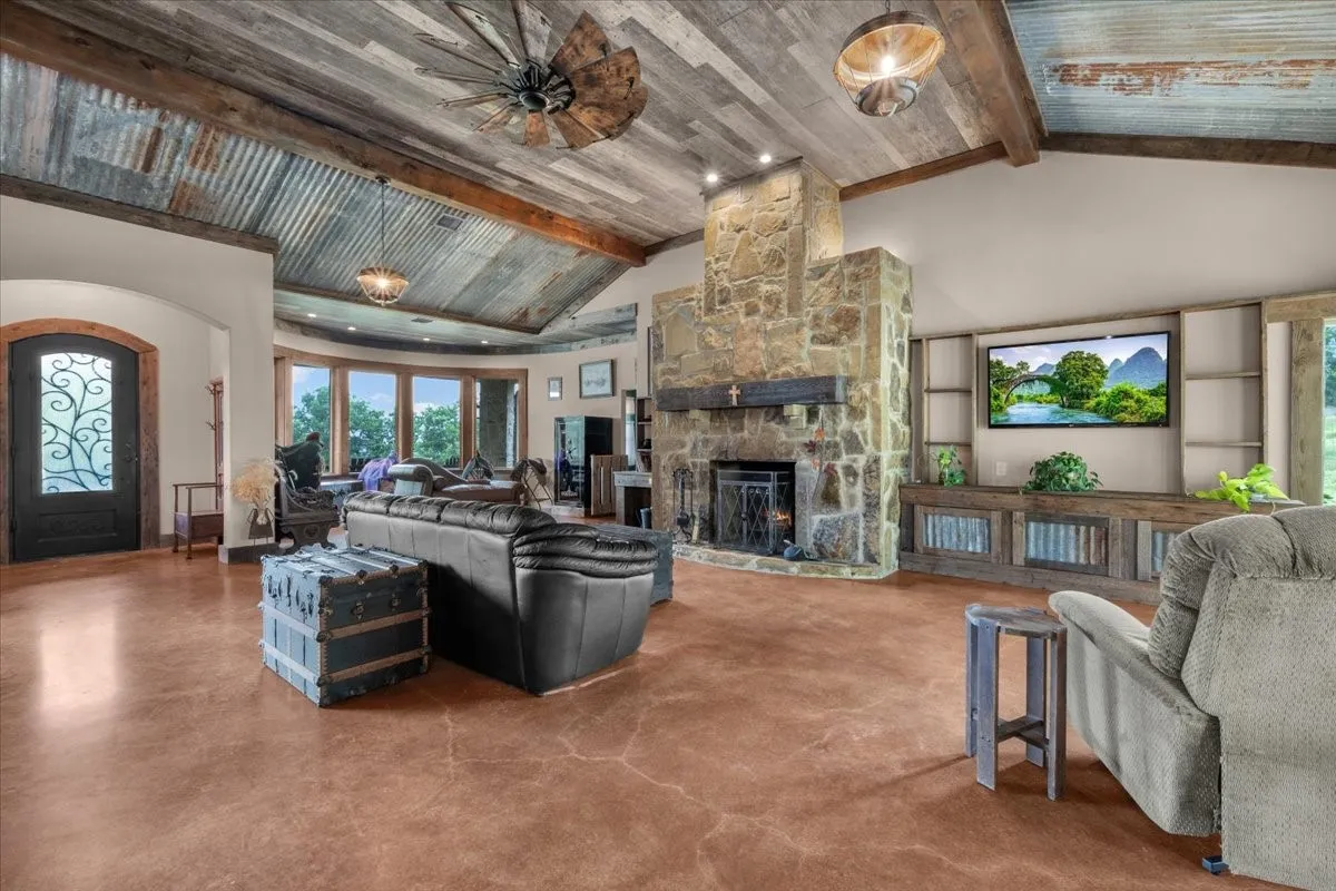 Spacious living area featuring a vaulted ceiling with wood paneling and corrugated metal accents, a floor-to-ceiling stone fireplace with a wood mantle, and an entertainment center with built-in shelving and rustic wood cabinetry