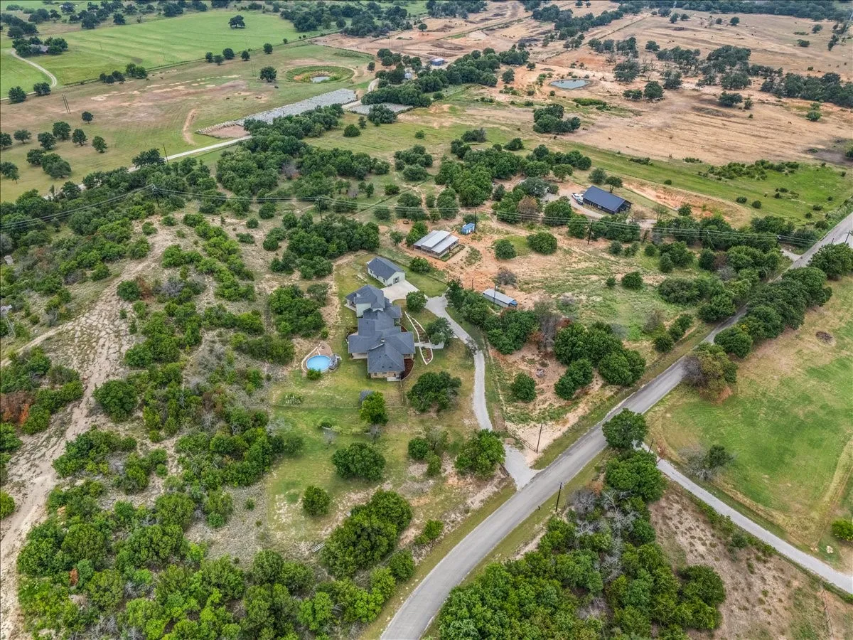 Expansive property featuring a main residence with a dark roof, a distinct blue-tiled swimming pool, and multiple additional structures, all surrounded by mature green foliage and open land