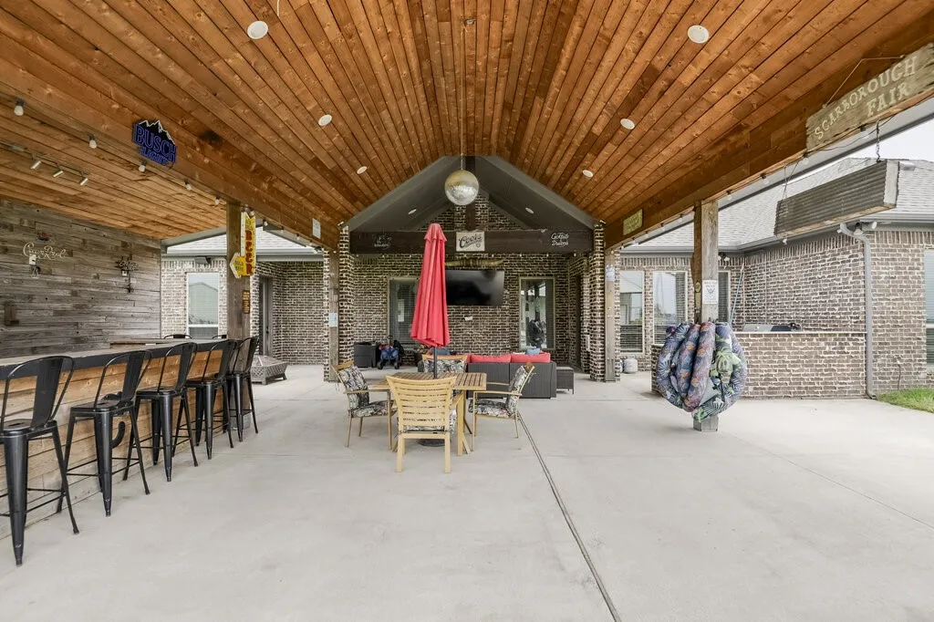 Covered outdoor living area featuring a wood-panel ceiling with recessed lighting, an expansive concrete patio, a built-in bar with a rustic wood-plank facade, a brick exterior, and an overhead disco ball fixture