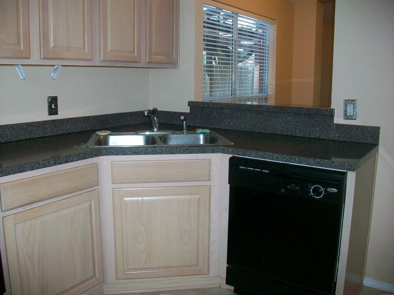 Kitchen workstation featuring a double-basin stainless steel sink, dark speckled countertop, light wood cabinetry, and a built-in black dishwasher