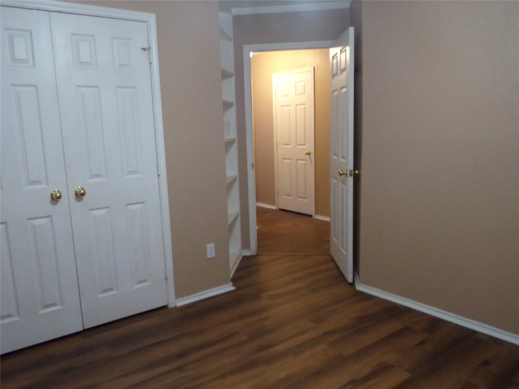 Wood-finish flooring throughout, featuring a double-door closet with gold-toned hardware, white baseboards, and built-in shelving