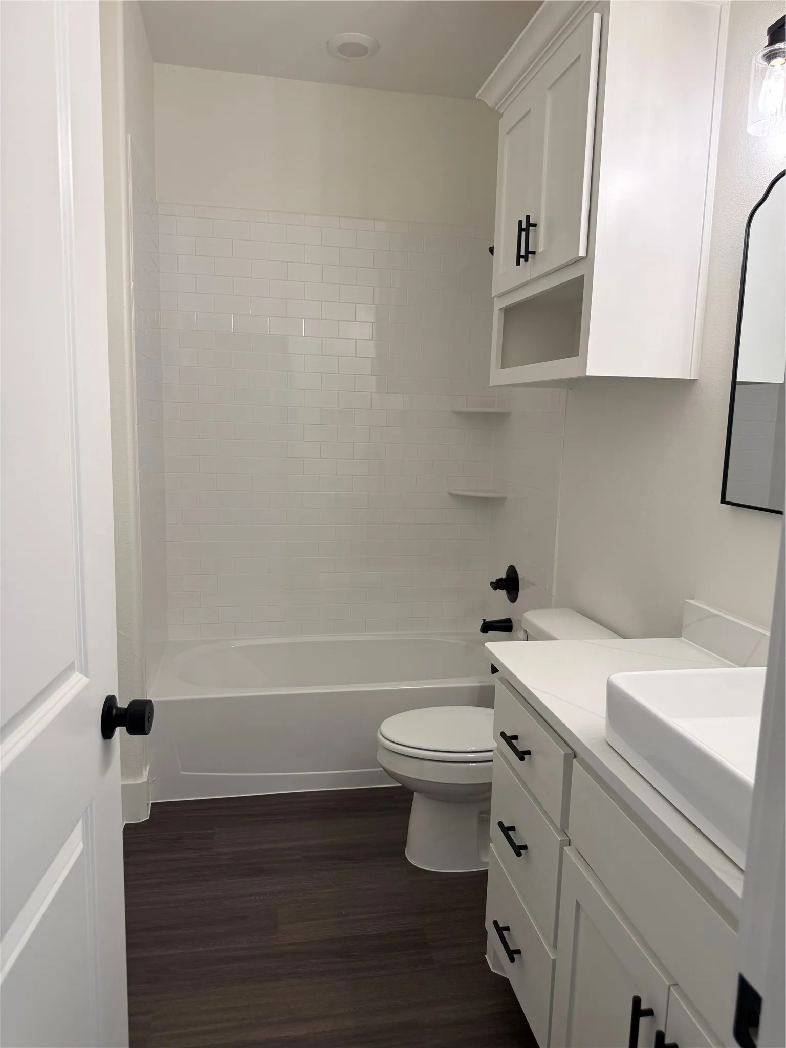 Contemporary bathroom featuring dark wood-finish flooring, a white vanity with vessel sink and white countertop, white shaker cabinetry with matte black hardware, a white bathtub with subway tile surround, and matte black plumbing fixtures