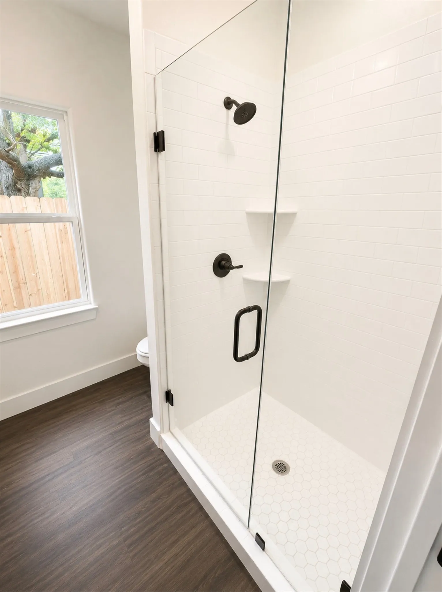 Modern shower enclosure featuring a frameless glass door, matte black fixtures, white subway tile surround, white hexagonal mosaic floor, and built-in shelving