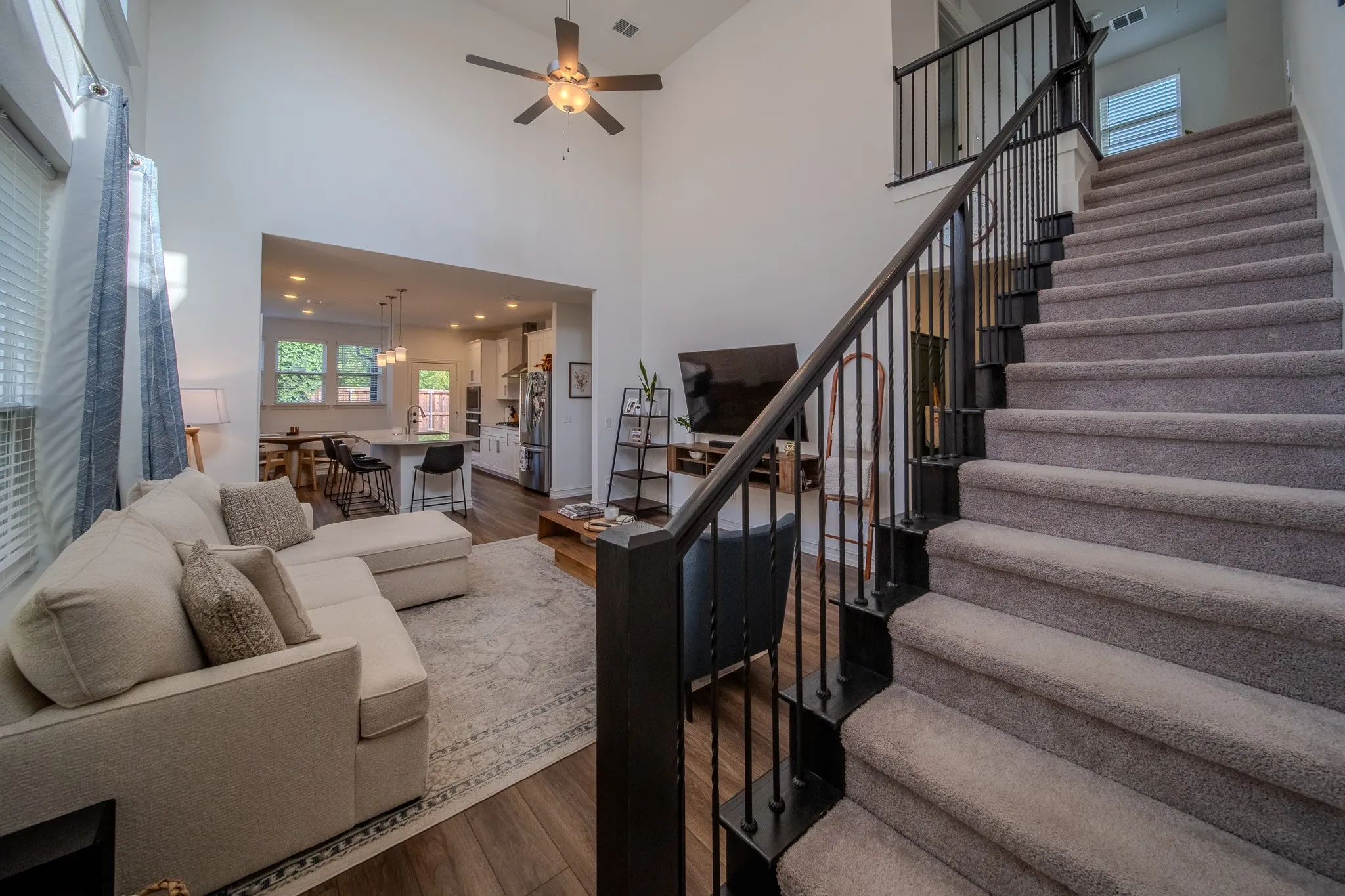Two-story living area featuring a vaulted ceiling with a ceiling fan, wood-finish flooring, and a carpeted staircase with dark metal balusters