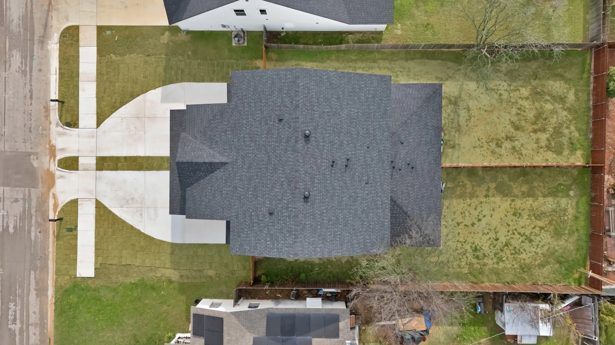 Aerial perspective showcasing a newly constructed property with a dark shingle roof, expansive concrete driveway, and established green lawn
