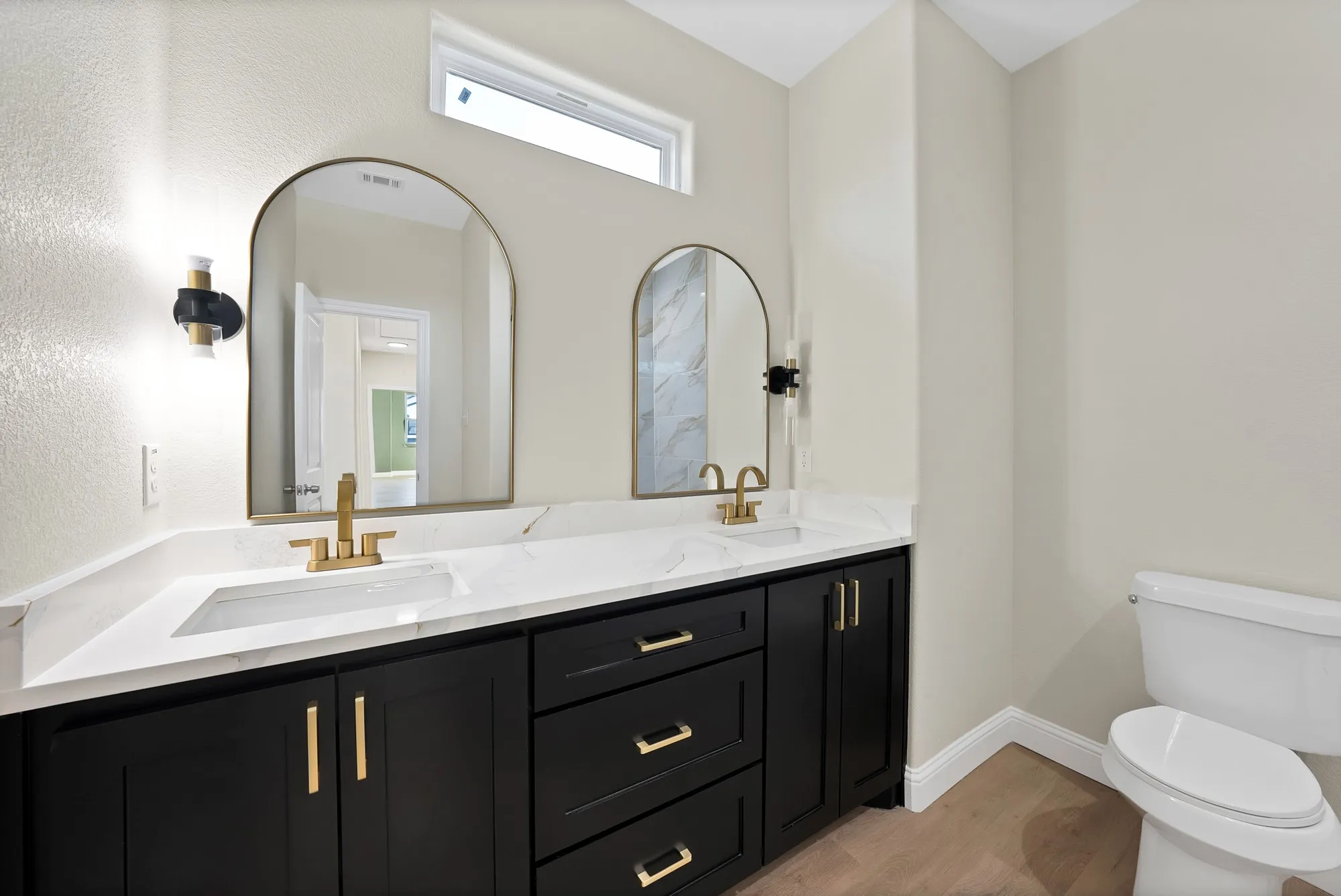 Double vanity with a white countertop and integrated sinks, dark cabinetry with gold-tone hardware, and arched mirrors with gold-tone frames