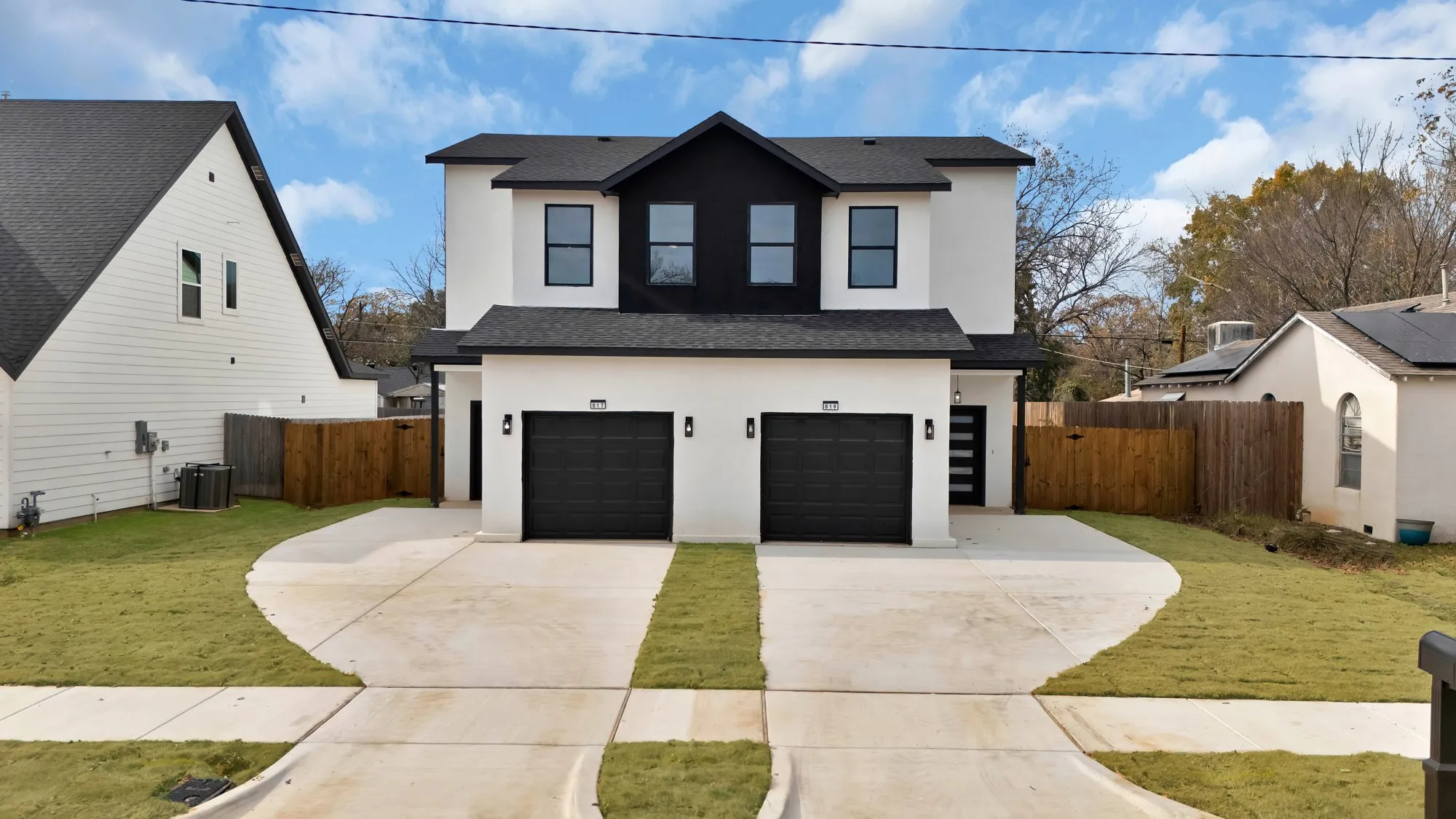 Modern duplex exterior featuring a contrasting black and white facade, dual black garage doors, two separate concrete driveways, and distinct entryways
