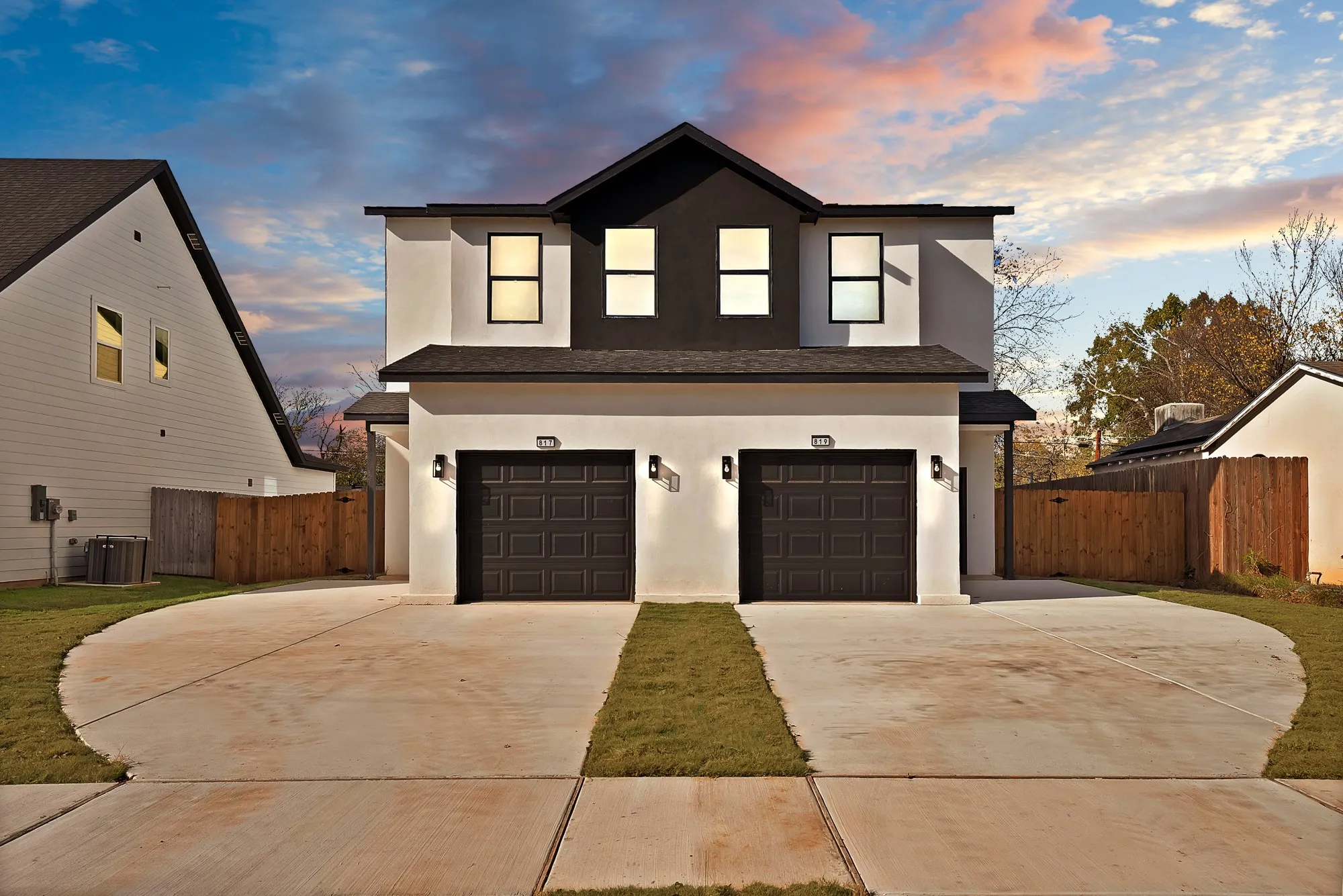 Contemporary duplex exterior featuring a contrasting black and white facade, dual garage doors, individual driveways, and modern exterior lighting