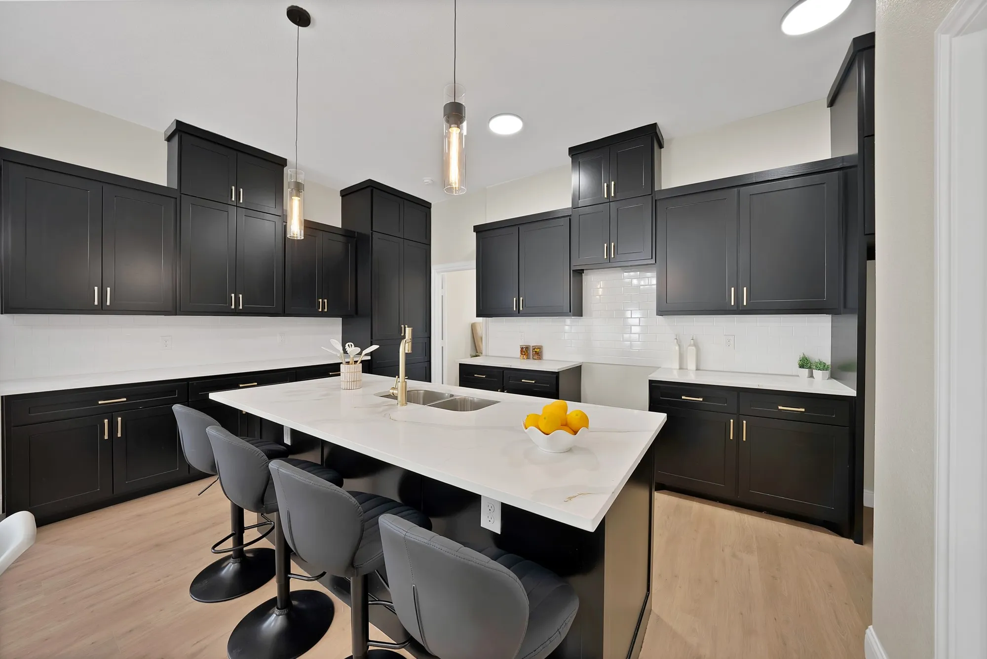 Kitchen featuring extensive dark cabinetry, white subway tile backsplash, light-toned wood-finish flooring, a large island with a built-in sink, and pendant lighting fixtures