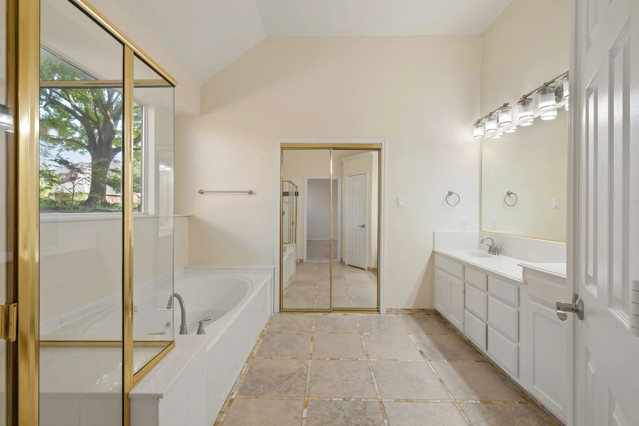 Spacious bathroom featuring a built-in tub with gold-framed glass enclosure, vaulted ceiling, dual sink vanity with white cabinetry, mirrored closet doors, and large format floor tile