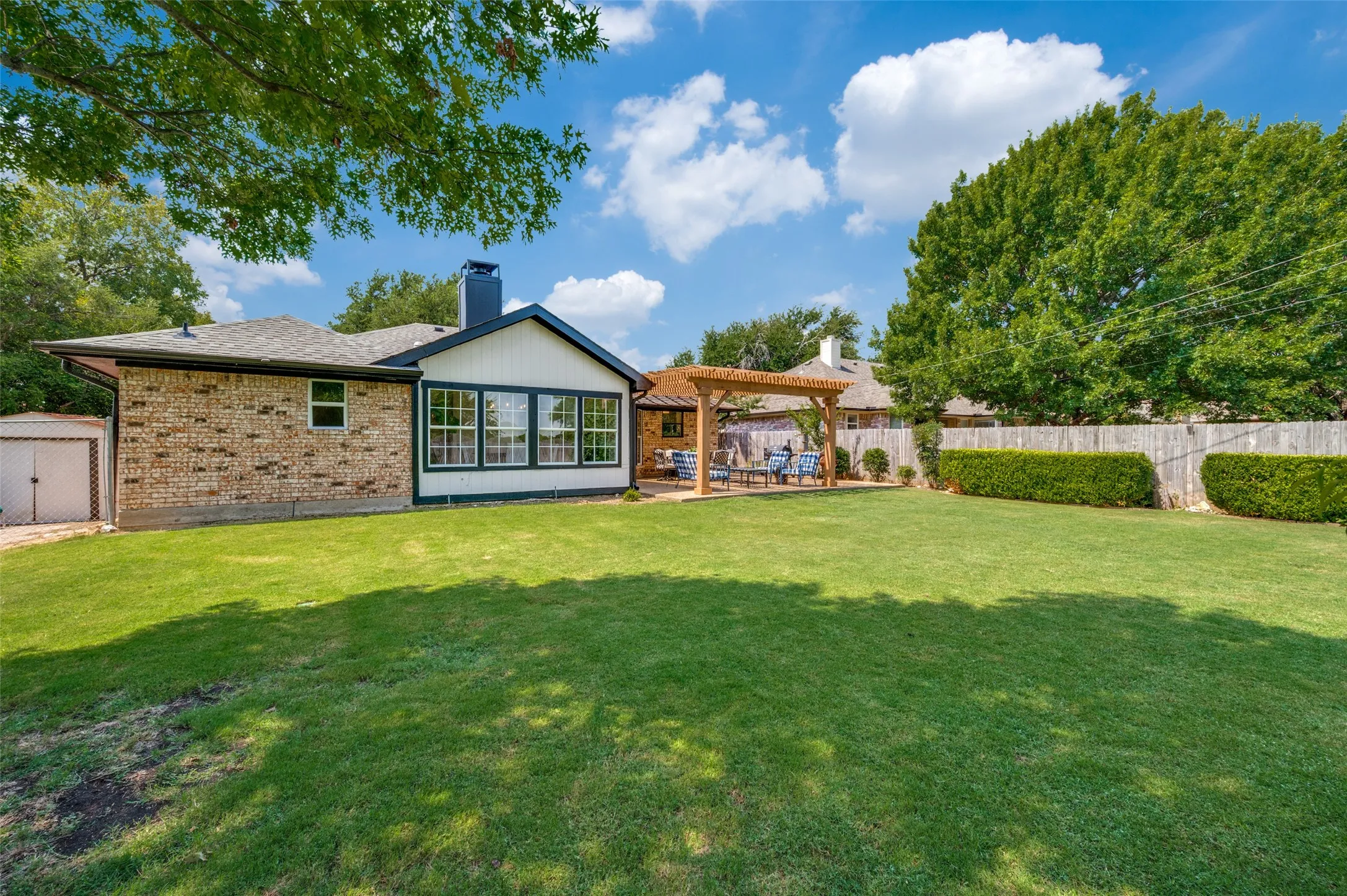 Backyard has lots of play space, a brick and white siding exterior, multiple windows, a dedicated patio area with a wooden pergola, and with a storage building tucked away from the main views