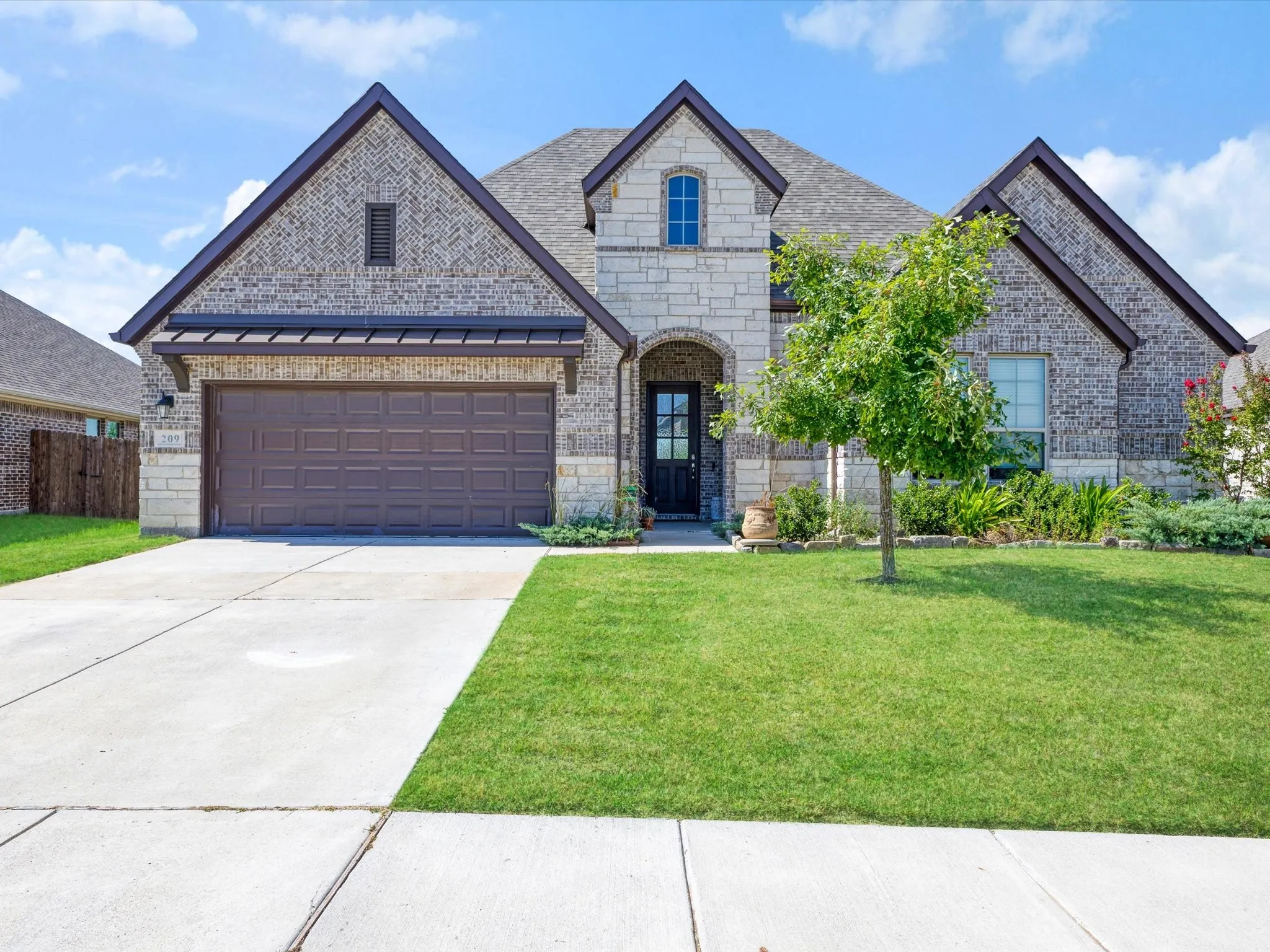 Exterior featuring a mixed material façade with brick and stone accents, a two-car garage with a brown panel door, an arched entry, a concrete driveway, and a front lawn with mature landscaping