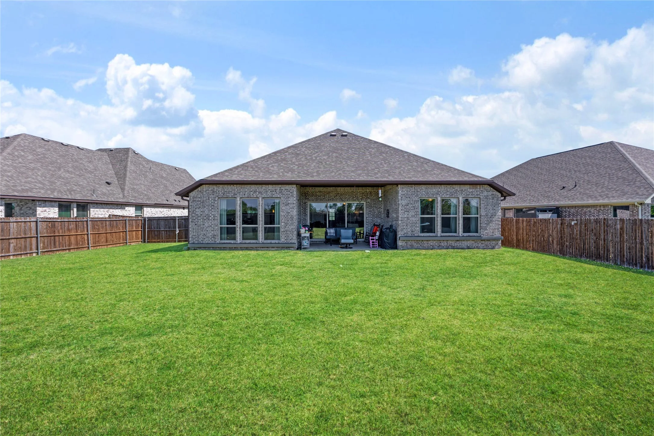 Expansive backyard featuring a manicured lawn, brick exterior, covered patio, multiple large windows, and a wood privacy fence