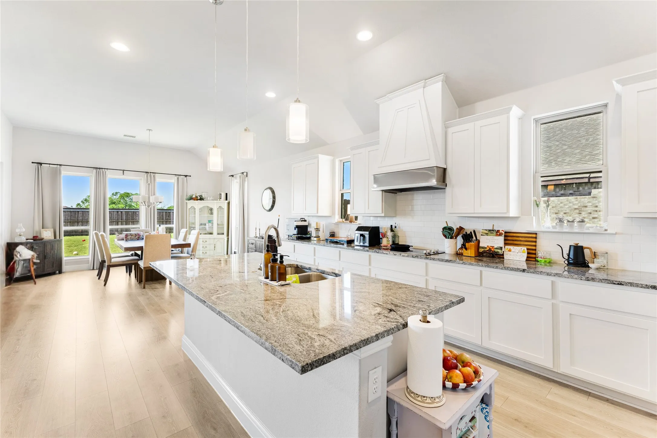 Bright kitchen featuring a large central island with an undermount sink, granite countertops, white cabinetry, a built-in range hood, and wood-finish flooring