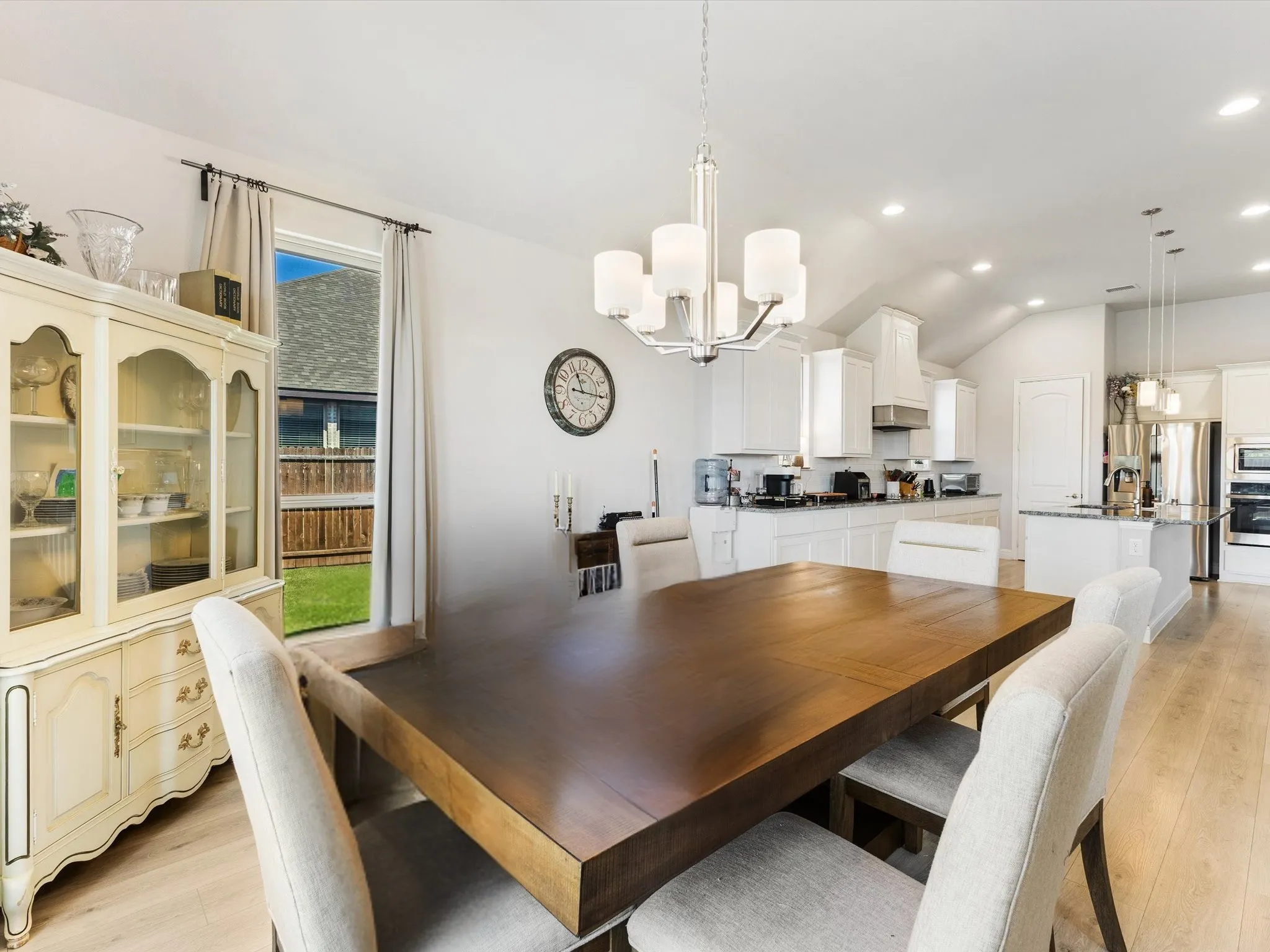 Open-concept dining area featuring light wood-finish flooring, a contemporary chandelier, vaulted ceilings, and a window with backyard views