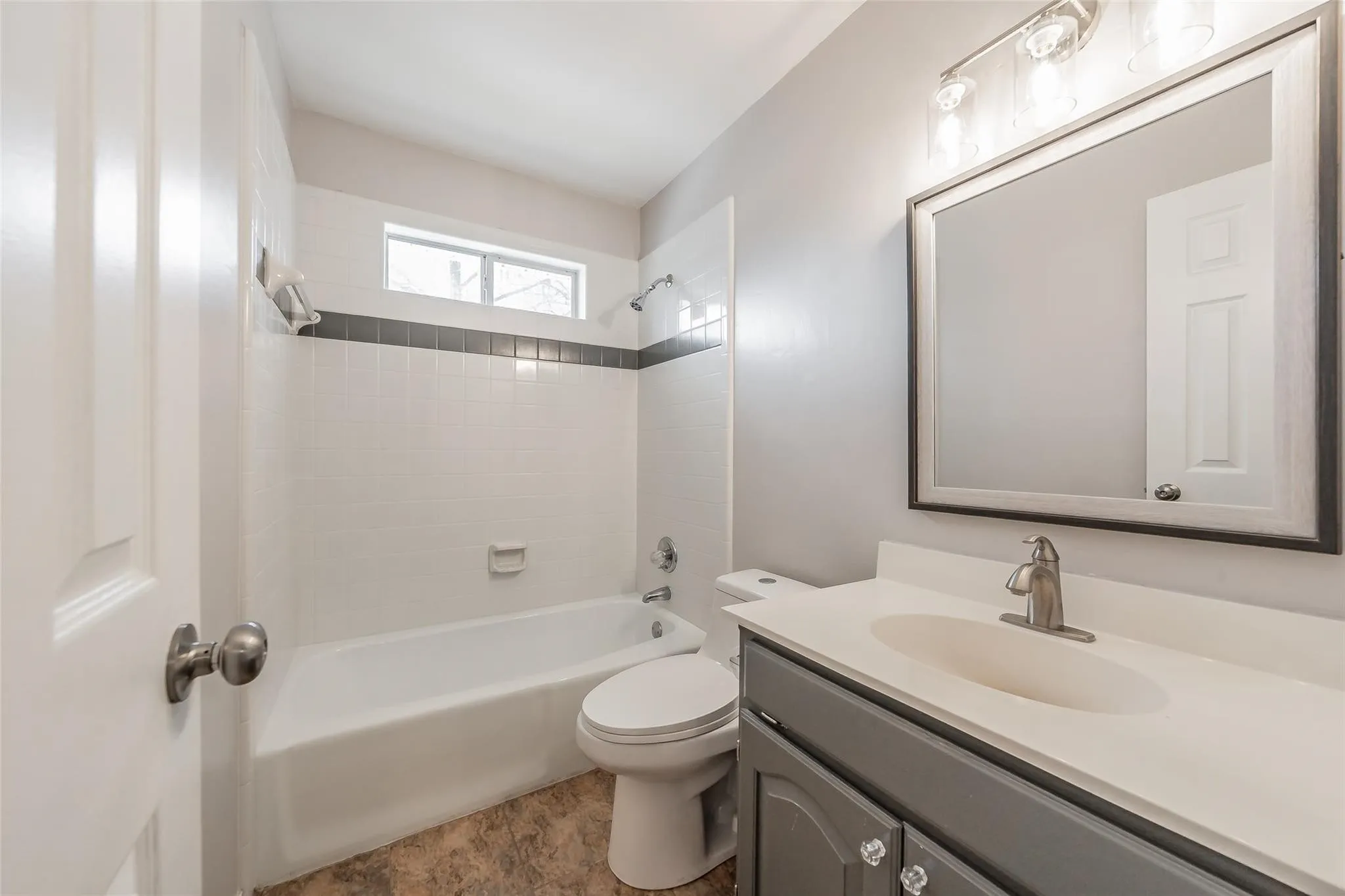 Bathroom featuring a white tile shower surround with a dark accent band, a built-in tub, and a vanity with a white countertop and gray cabinetry