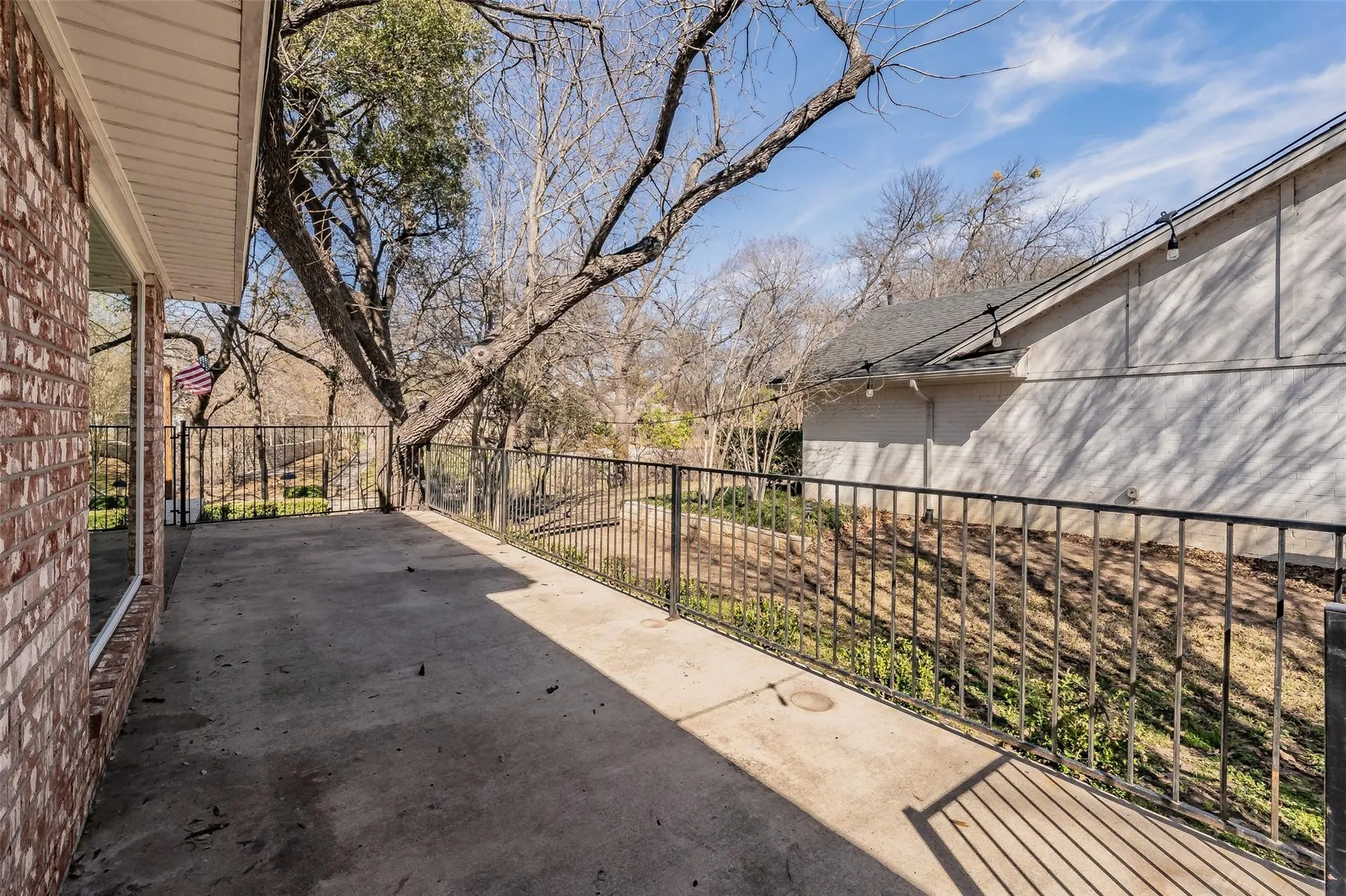 Concrete patio featuring a black wrought iron railing, brick exterior, and mature trees