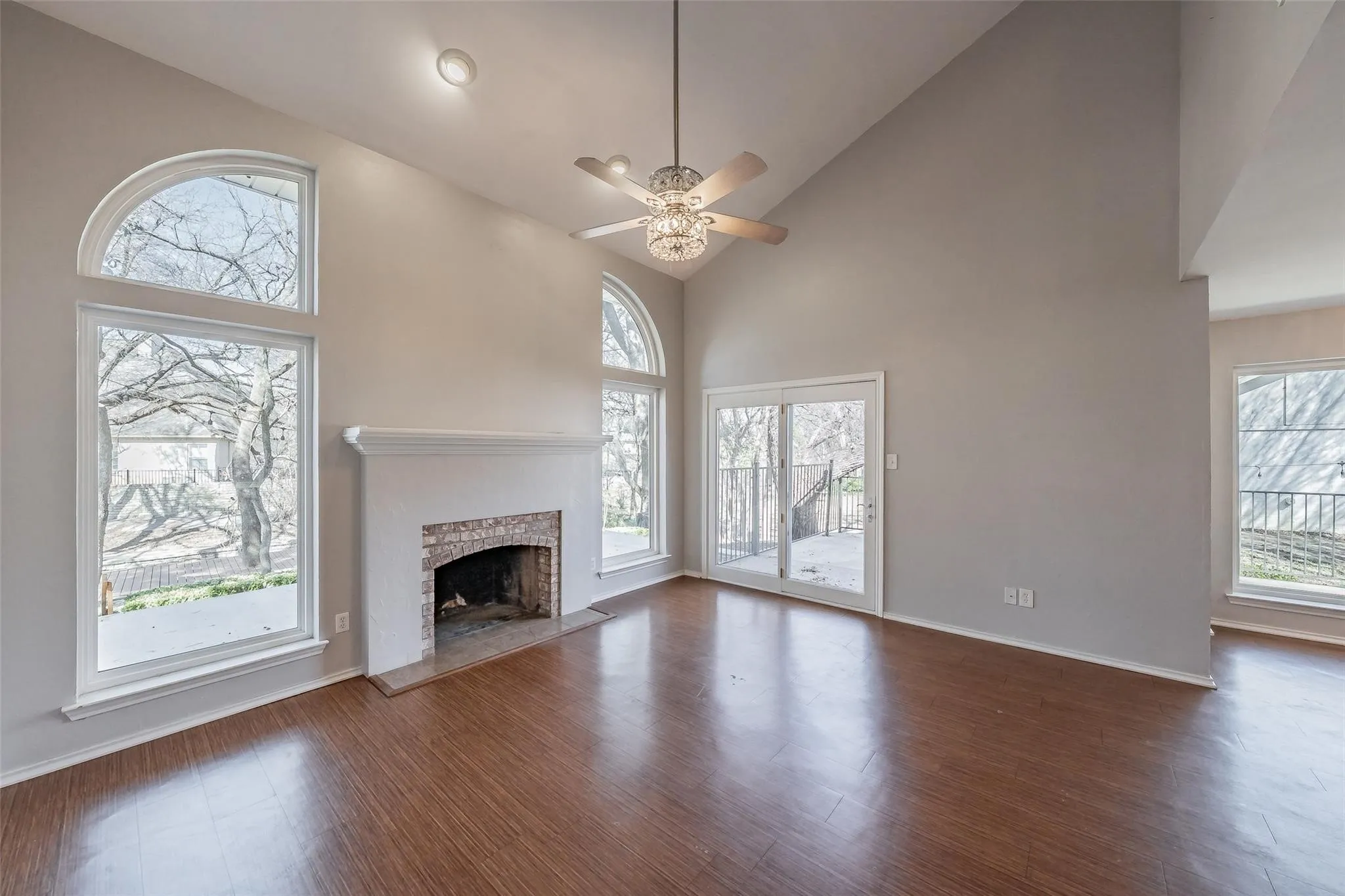 Spacious living area featuring high vaulted ceilings, wood-finish flooring, a brick fireplace with a white mantel, and multiple large windows with arch-top transoms