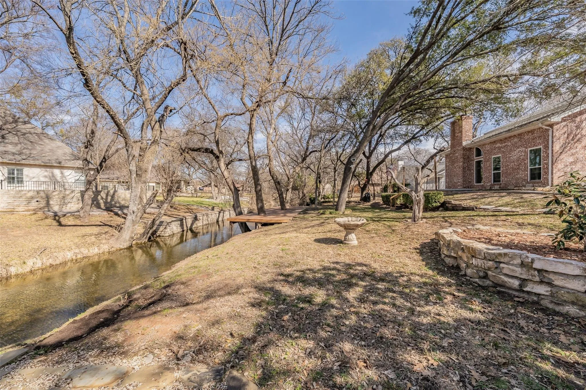 Expansive backyard featuring a natural water channel, a private wooden deck, mature trees, a stone bird bath, and a stone retaining wall
