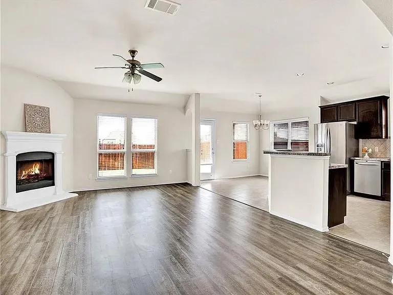 Open-concept living area featuring wood-finish flooring, a corner fireplace with white mantel, a ceiling fan, and multiple windows